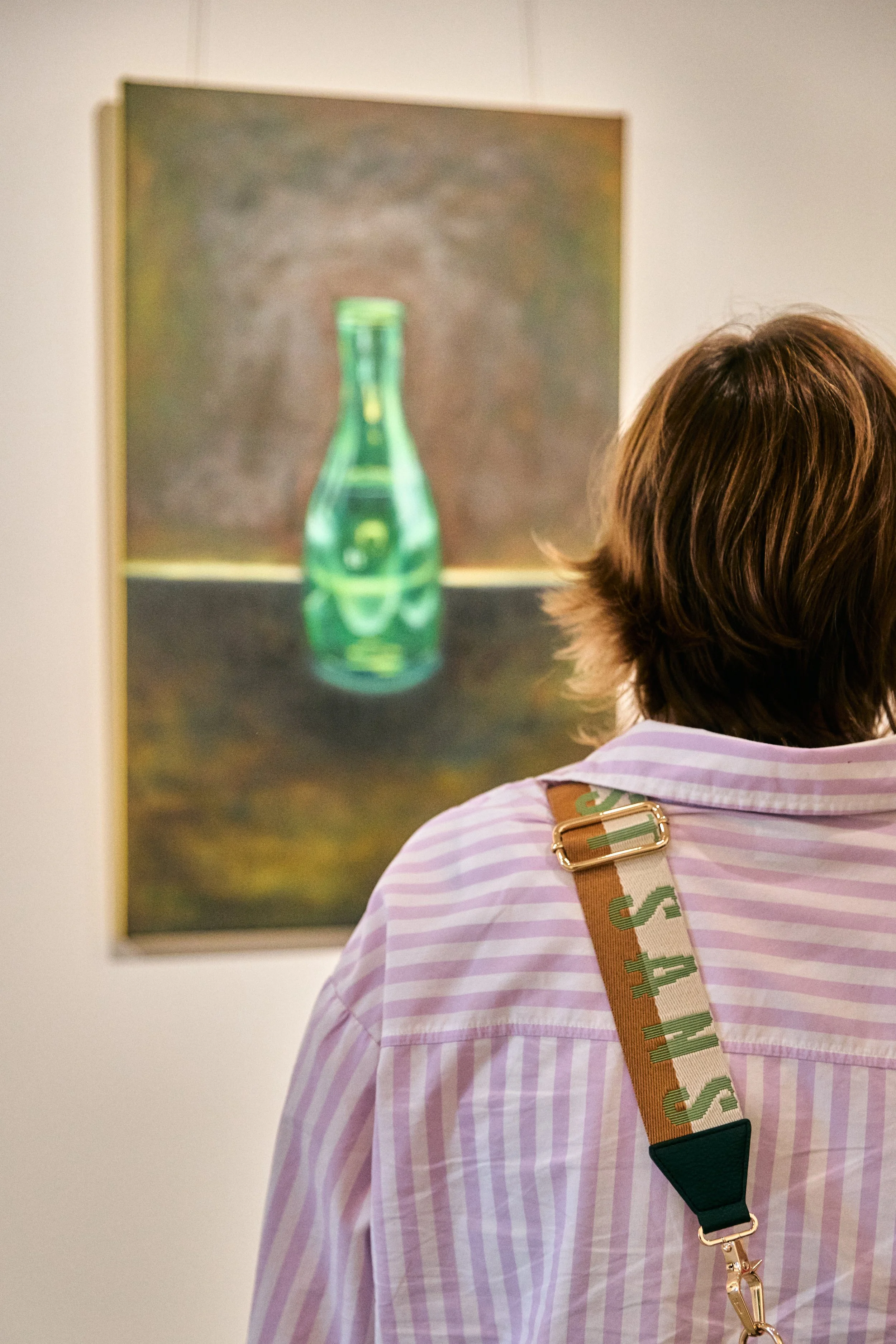 A person with brown hair wearing a pink and white striped shirt looking at a blurred painting of a green bottle on a gallery wall.