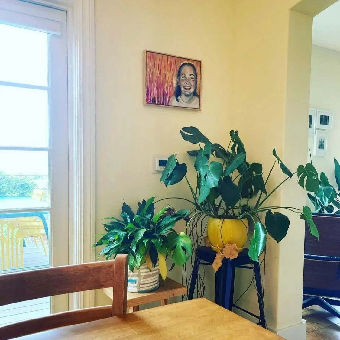 Interior of a home with potted plants on a small table and a stand, a framed portrait on the wall, a thermostat, and a sliding glass door leading outside with yellow chairs visible.