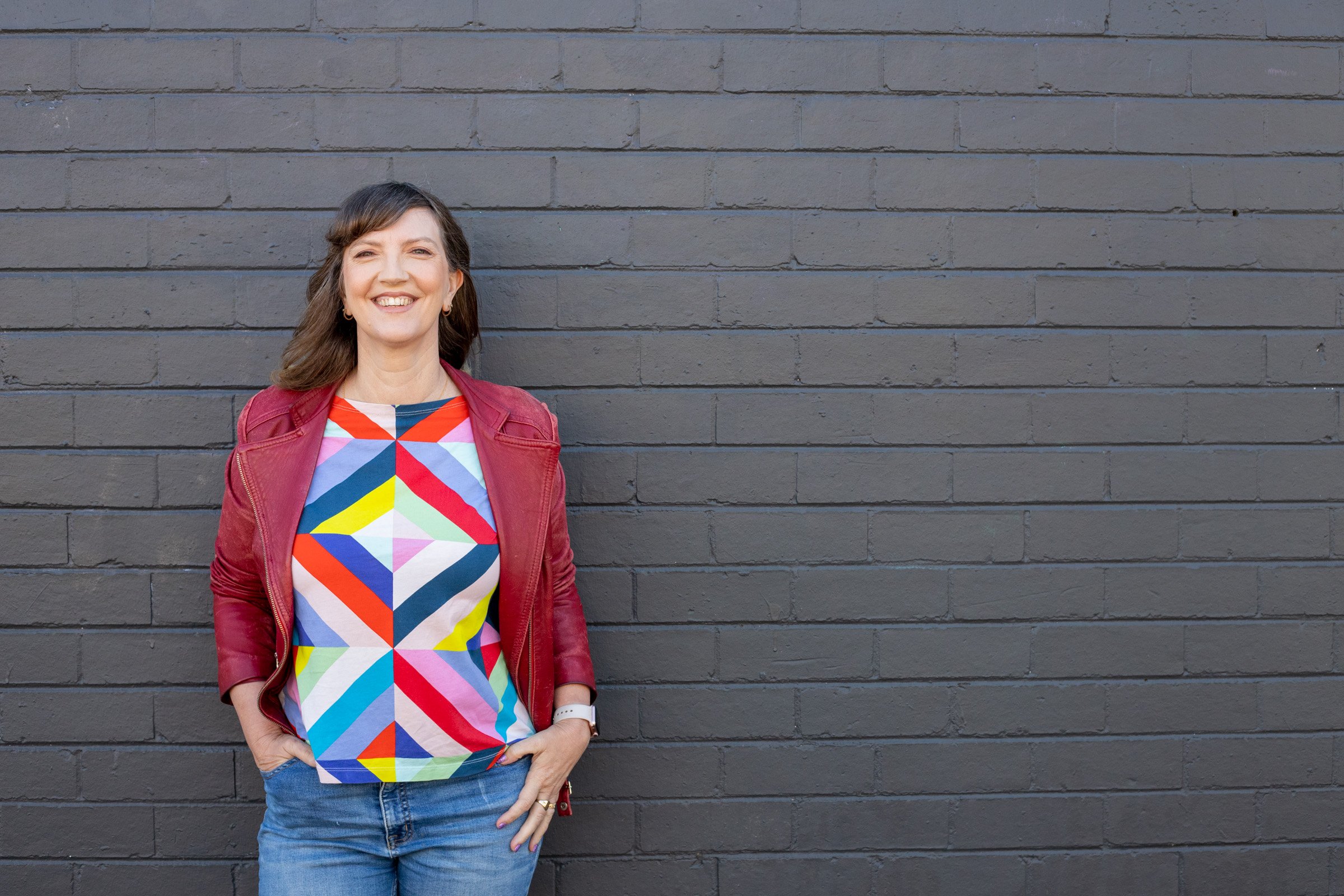 A woman with shoulder-length brown hair smiling, wearing a colorful geometric-patterned shirt, a red jacket, and blue jeans, standing against a dark gray brick wall.