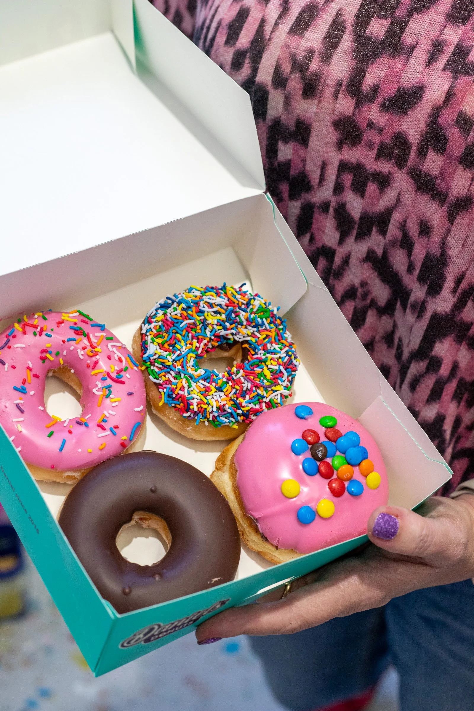 A box of assorted colorful donuts, including pink frosted with rainbow sprinkles, chocolate frosted, and pink frosted with multicolored round candies, held by a person with purple glittery nail polish.