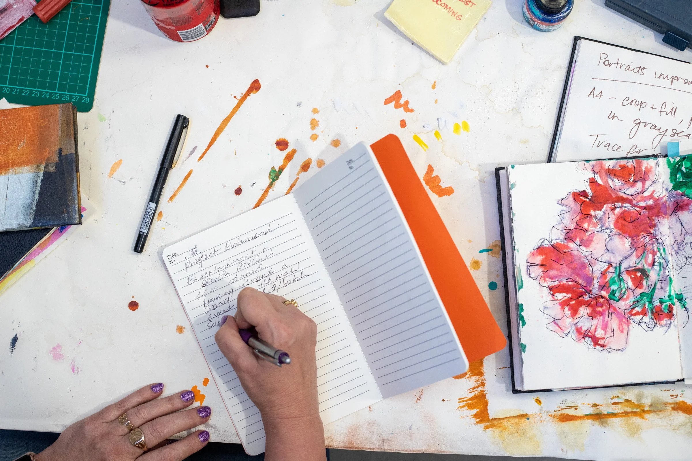 A person with purple glittery nails writing in a notebook with handwritten notes on a white desk with paint splatters, surrounded by art supplies, a black pen in hand, and colorful artwork and notebooks.