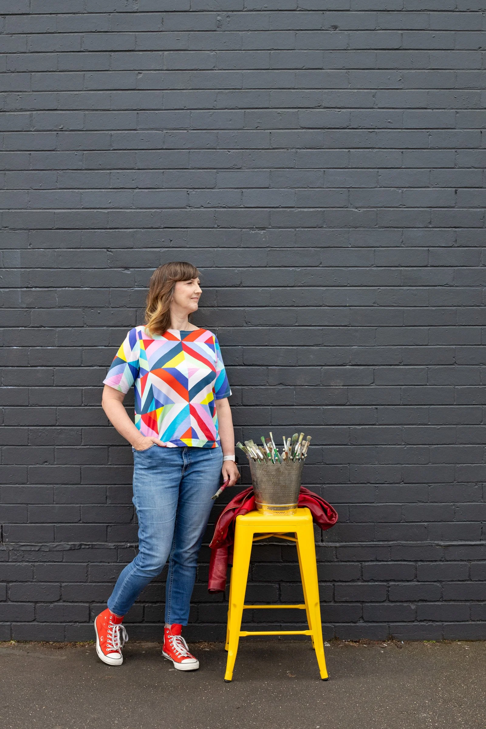 A woman with light brown hair in loose waves, wearing a colorful geometric pattern shirt, blue jeans, and red sneakers, standing against a dark gray brick wall. Next to her, there is a yellow stool with a metal container filled with paintbrushes, a red jacket draped over the stool, and a dark red bag underneath.