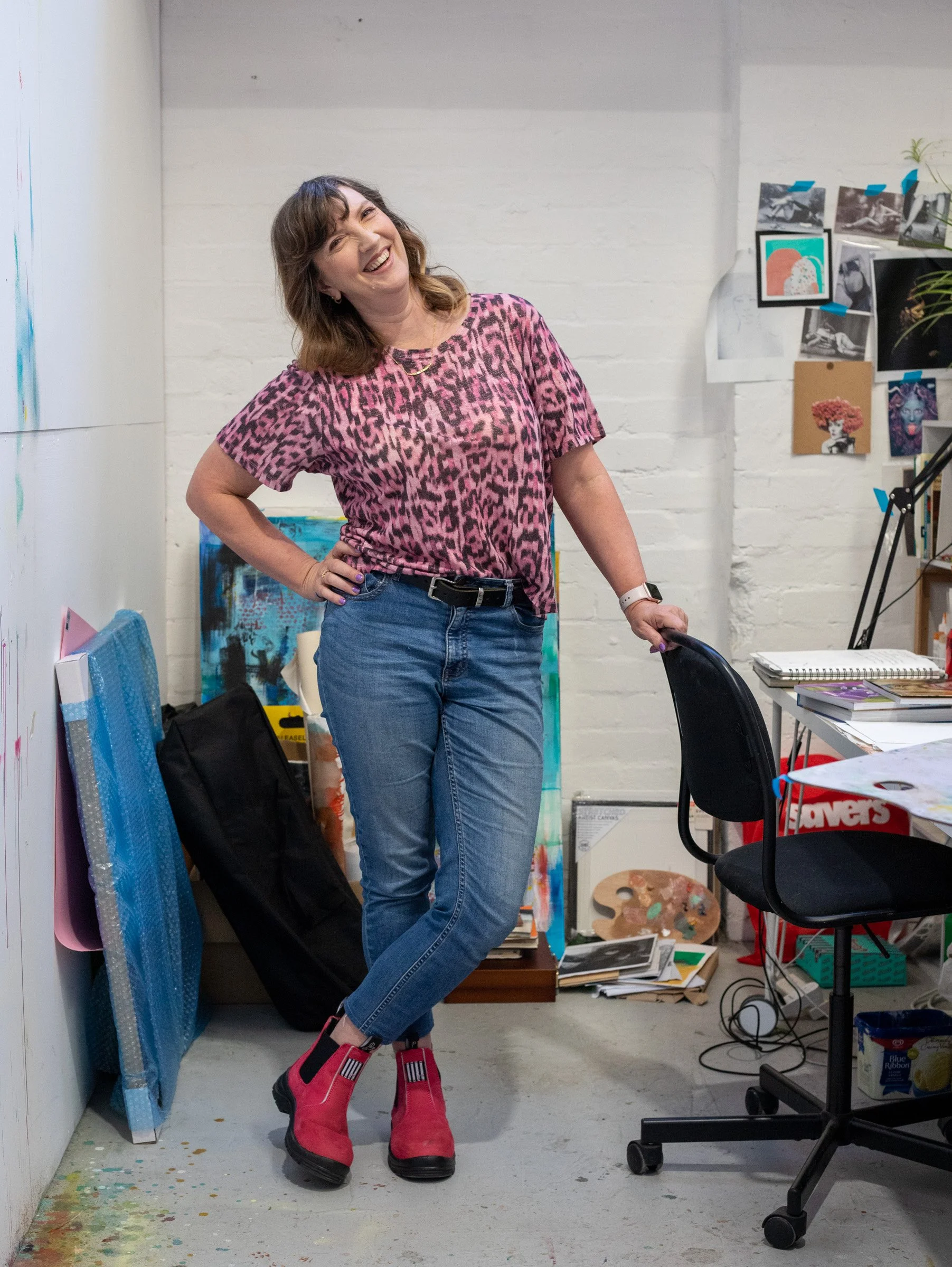 A woman standing in an art studio, laughing with her hand on her hip, wearing a pink leopard print shirt, blue jeans, and pink and black boots, surrounded by art supplies and artwork.