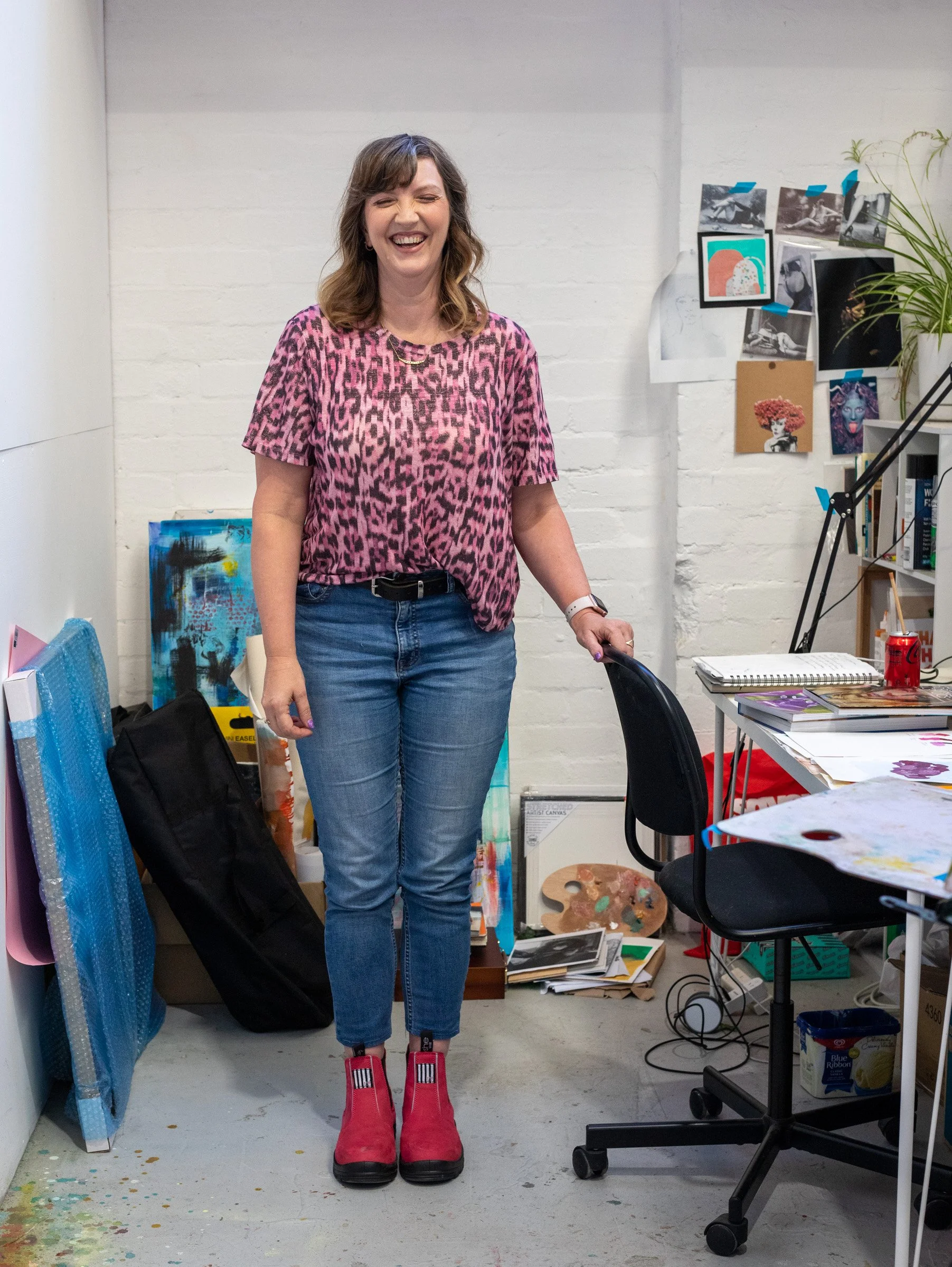 A woman with brown, shoulder-length hair wearing a pink animal print shirt, blue jeans, and pink boots, standing in an art studio with a white brick wall, art supplies, and artworks in the background, smiling with eyes closed.