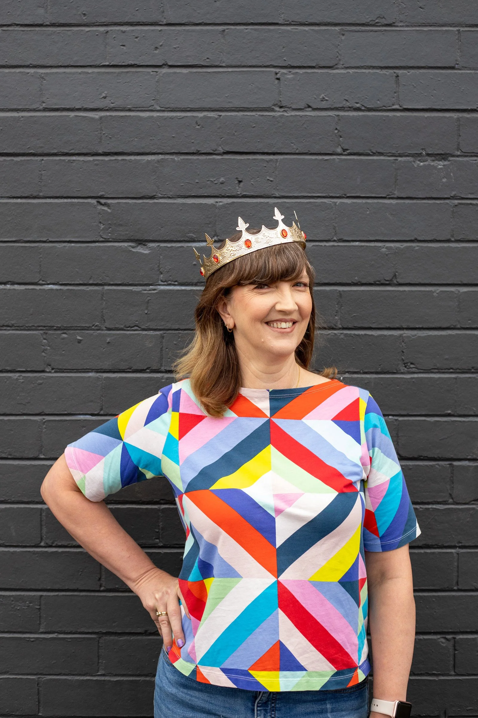 A woman wearing a colorful geometric patterned top, a gold crown with red gems, and blue jeans standing against a dark gray brick wall.