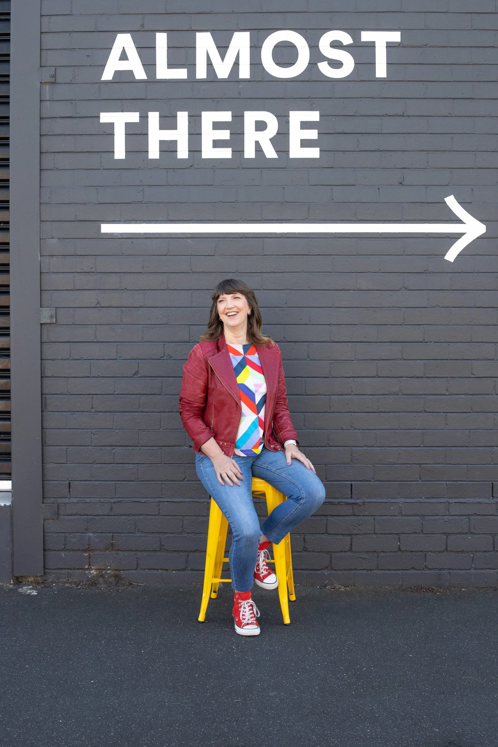 A woman sitting on a yellow stool in front of a dark gray brick wall with a large sign that reads 'Almost There' with an arrow pointing to the right. She is smiling, wearing a colorful shirt, a red jacket, blue jeans, and red sneakers.