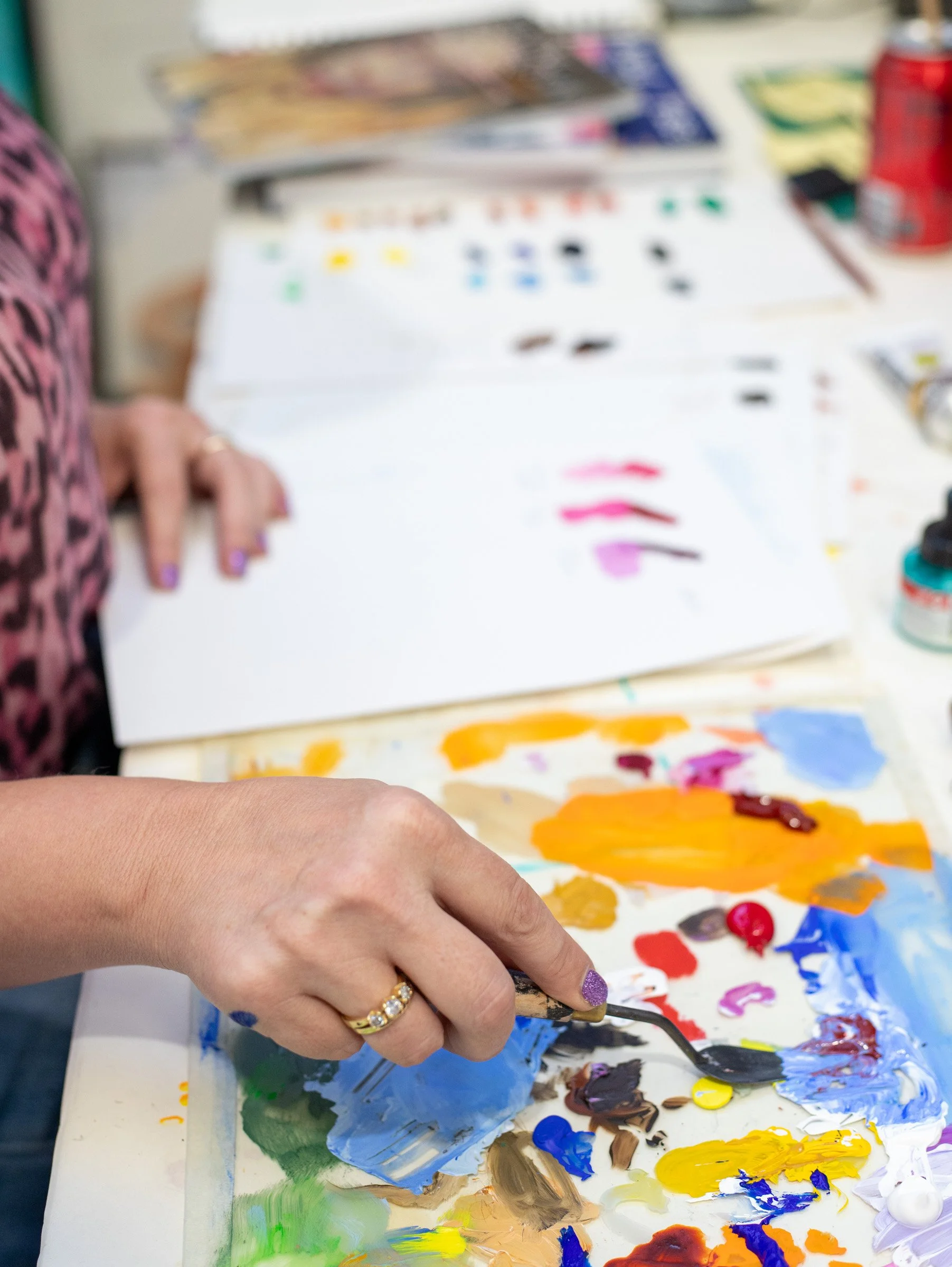 Close-up of a person mixing blue and yellow paint on a palette with a palette knife, with a multi-colored paint palette in the background.