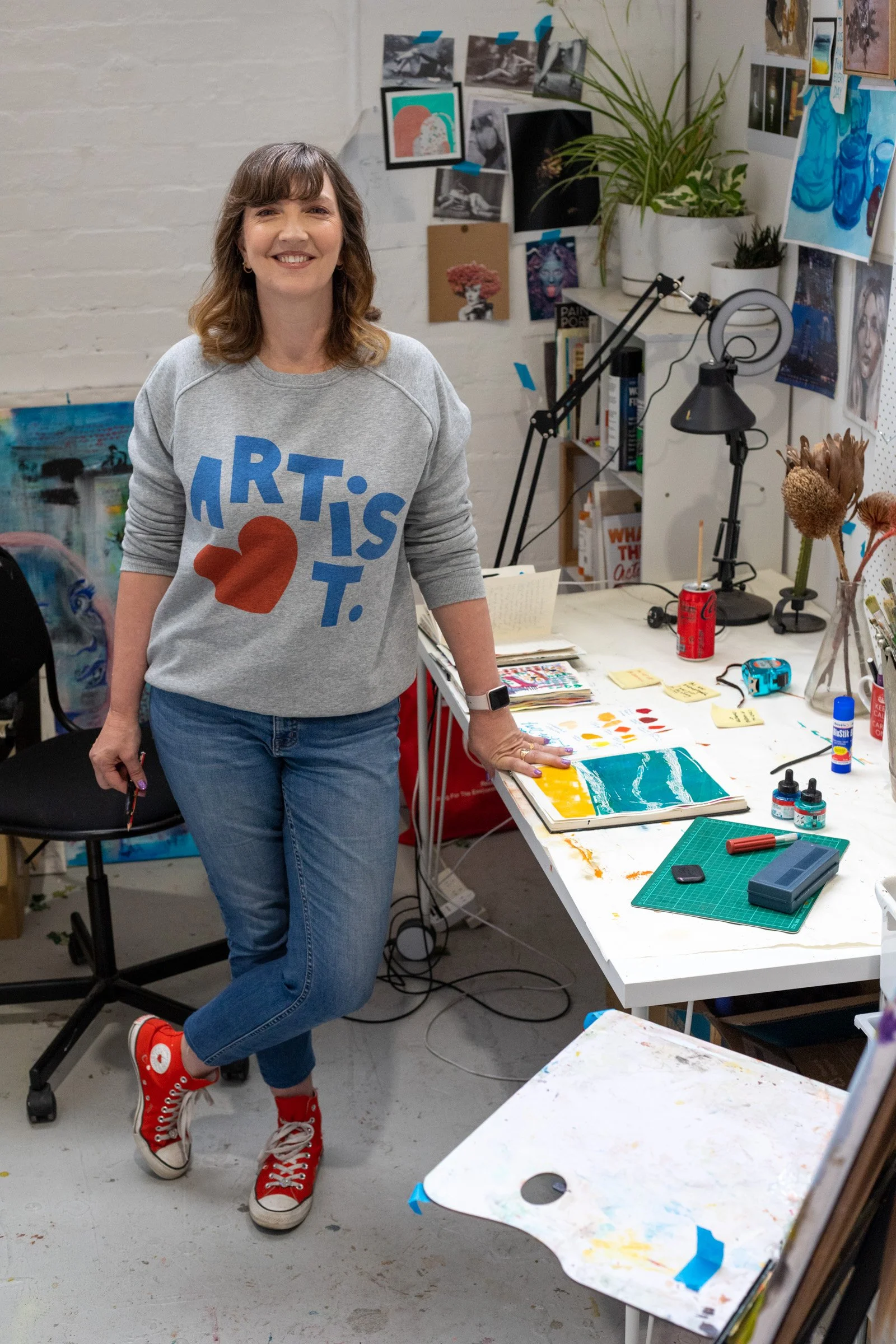 A woman with shoulder-length brown hair and a light gray sweatshirt with blue and red graphics stands in an artist's studio. She is smiling and leaning on a white table covered with paintings, art supplies, and a green cutting mat. The background shows shelves with books, potted plants, and artwork pinned to the walls.