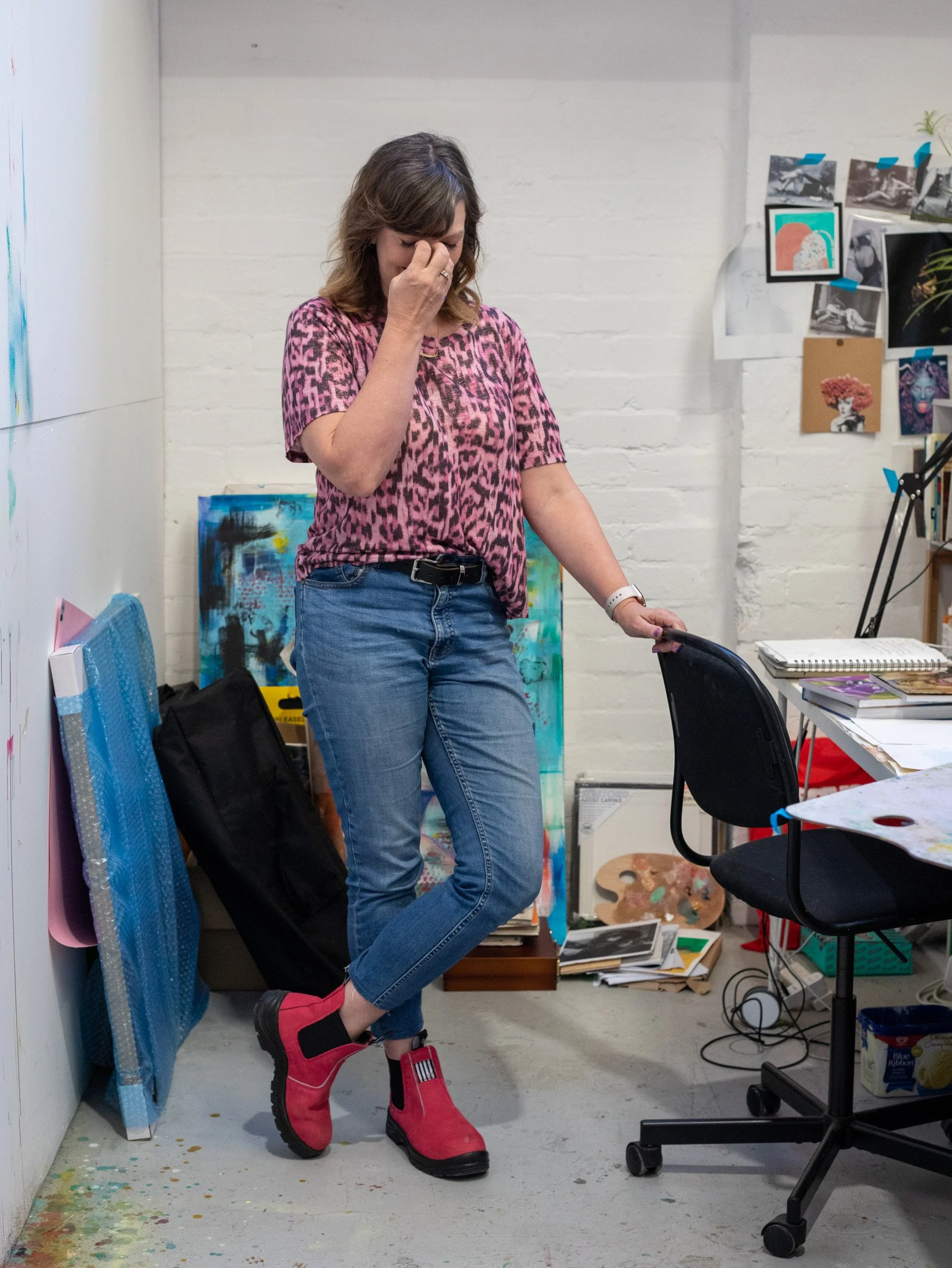 A woman stands in an art studio wearing pink and black leopard print shirt, blue jeans, and red rubber boots. She has short, wavy brown hair and is covering her face with one hand. The studio has artwork, supplies, and a white brick wall.