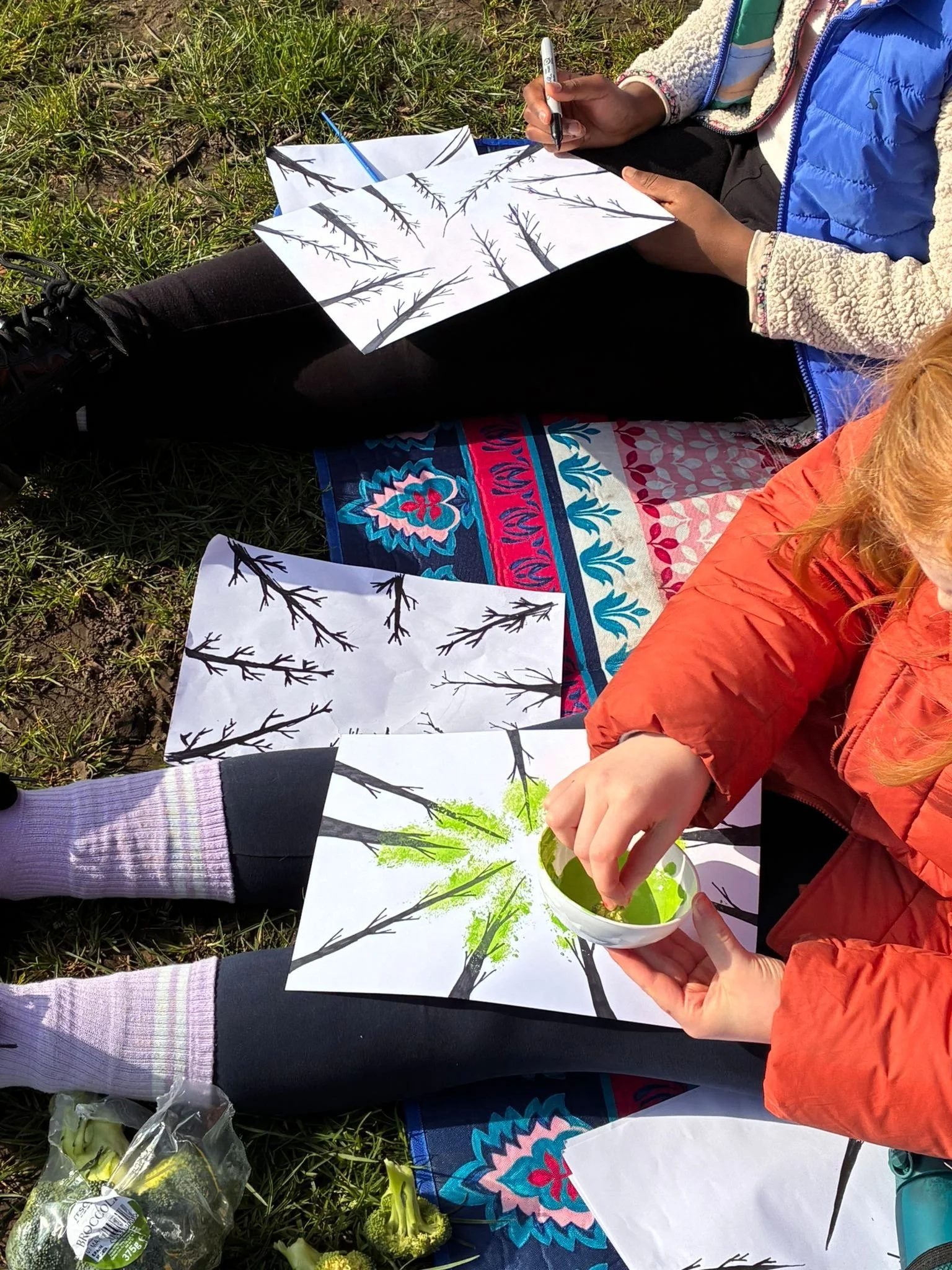 Two people sitting on a grassy ground painting tree silhouettes with green watercolor. They have papers with black outline tree sketches and are filling in with green. A bowl of green paint is held by one person. There are flowers and vegetables nearby.