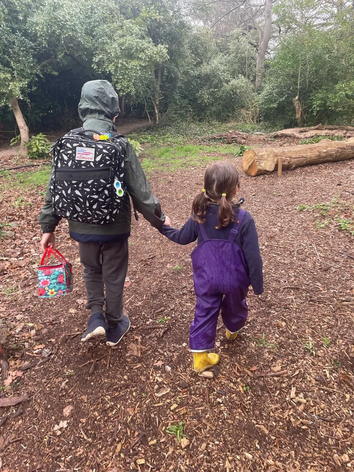 Two children holding hands while walking in a wooded area on a dirt trail, wearing rain jackets and backpacks.
