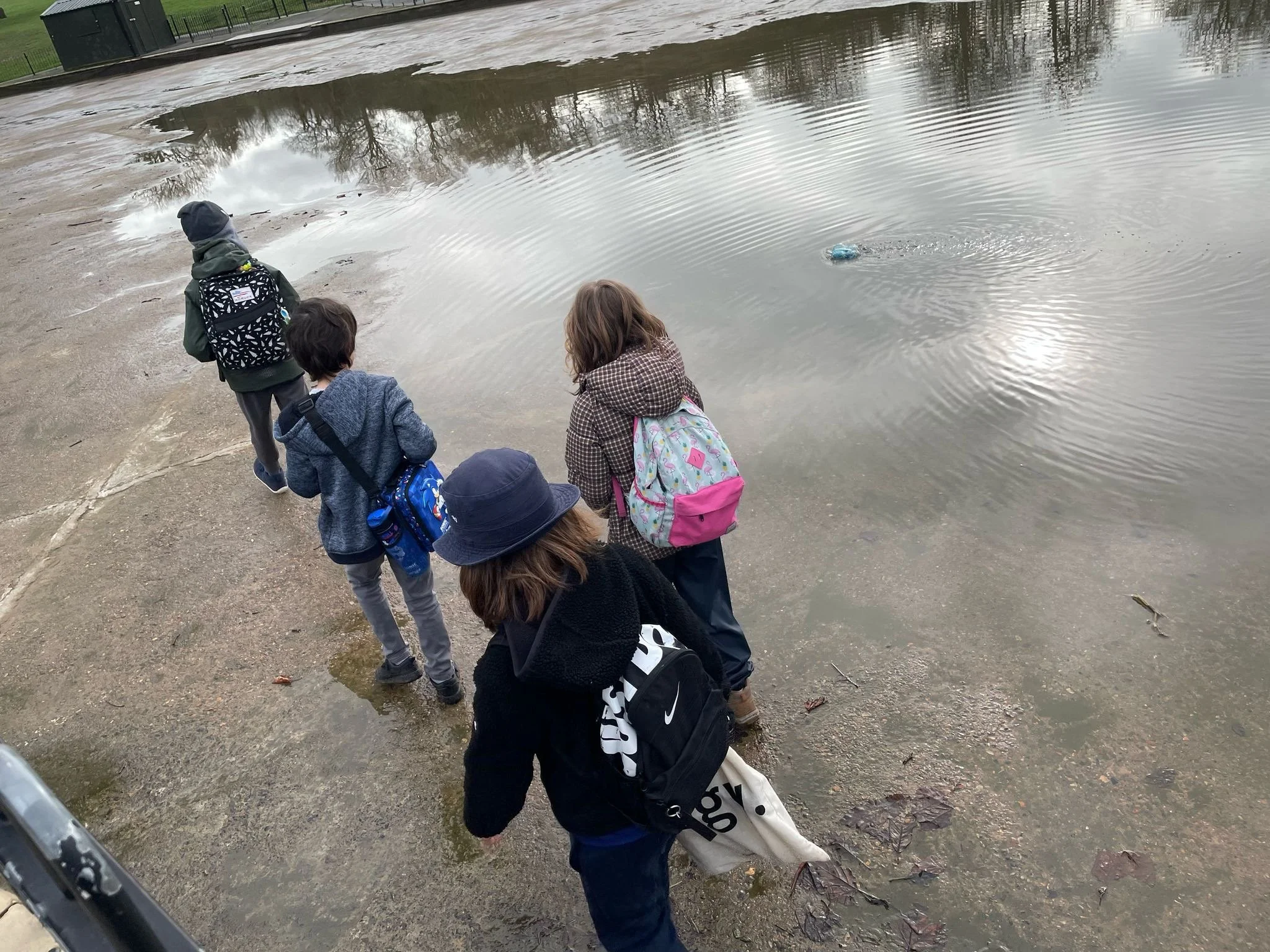 Five children with backpacks standing on a wet sidewalk near a pond, watching a small object floating on the water.