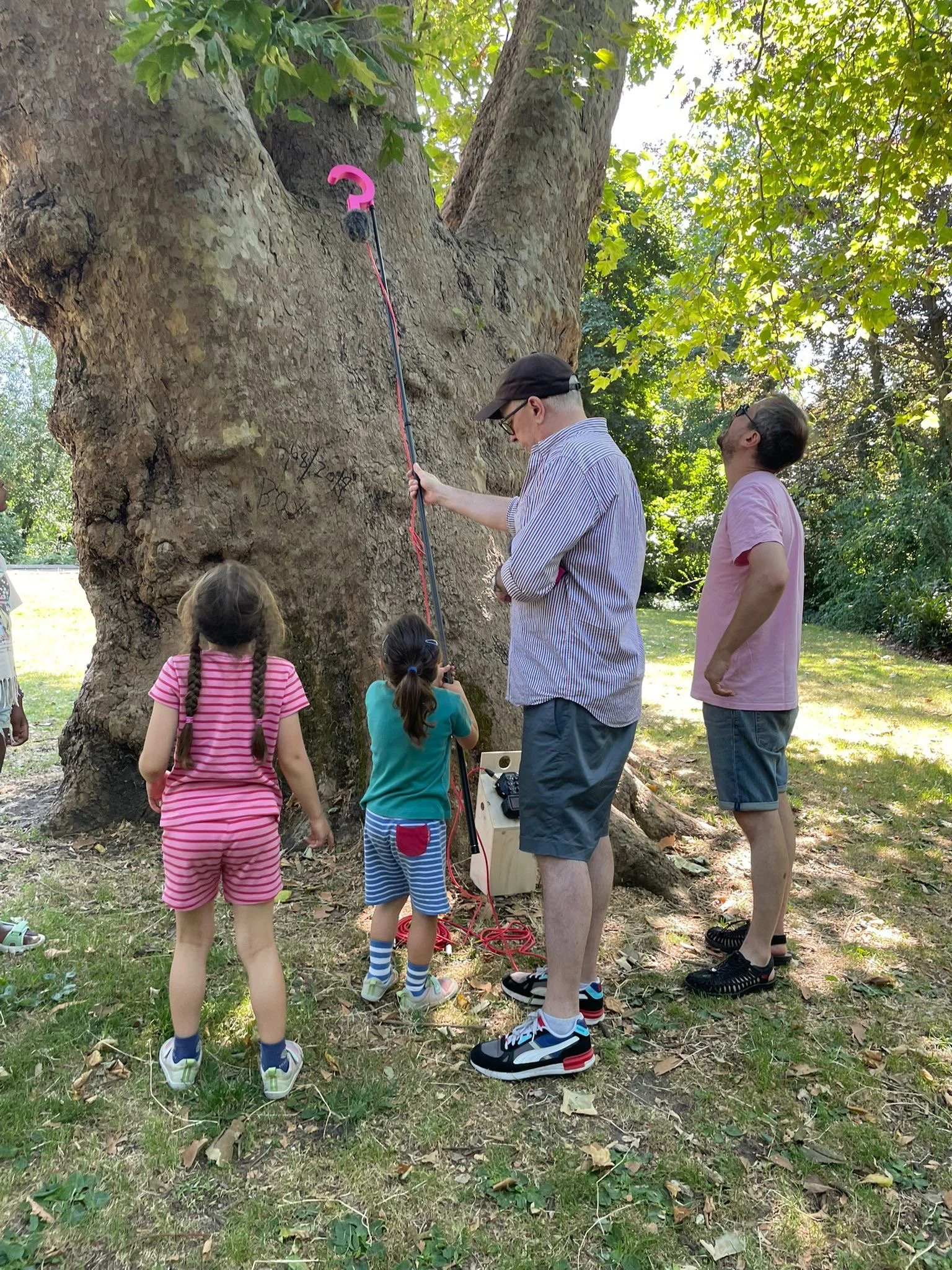 A group of people, including three children and two adults, are gathered around a large tree outdoors. One person is operating a measuring device or tool against the trunk of the tree, while the others watch. The scene is set in a sunny area with gre