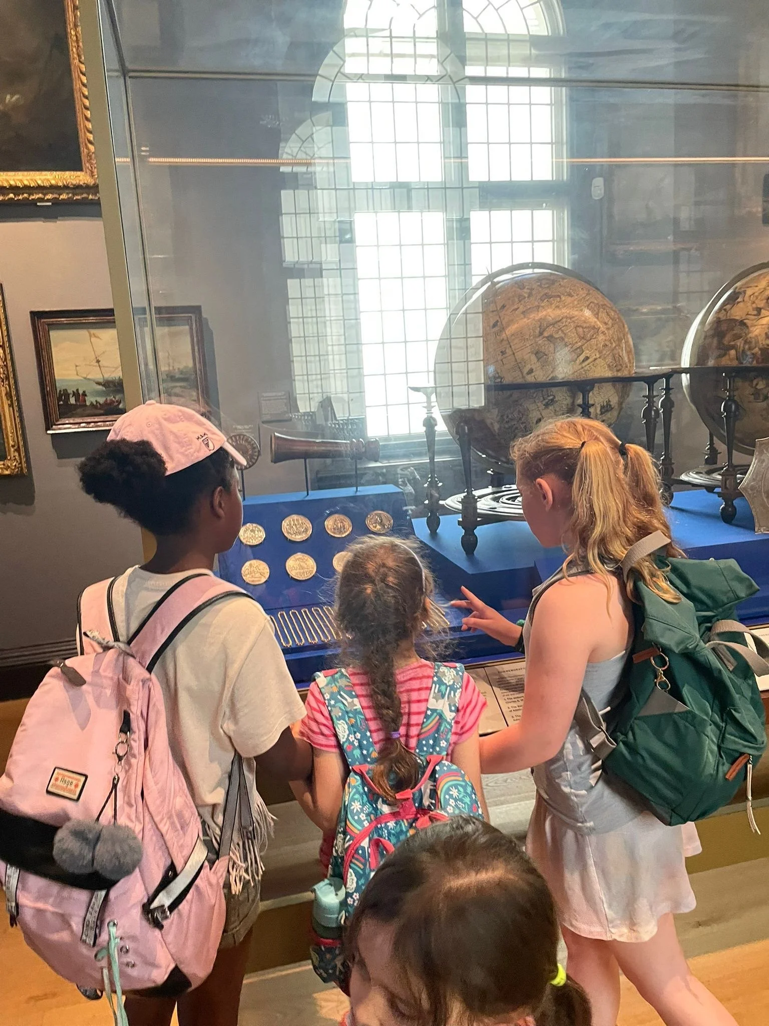 Children observing historical artifacts, including globes, coins, and a telescope, in a museum exhibit behind glass.