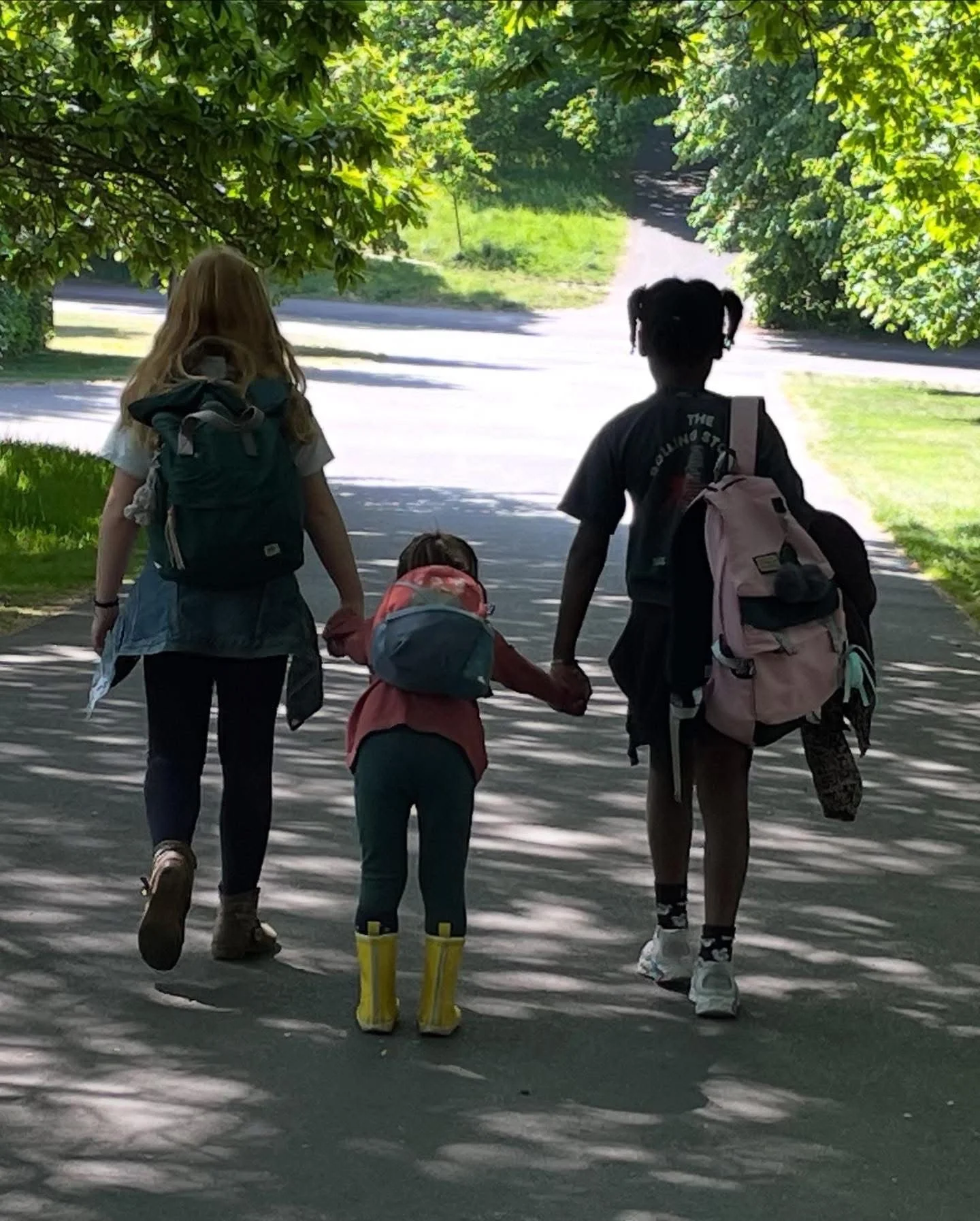 A woman, a young girl, and a teenage boy walking hand in hand down a shaded sidewalk on a sunny day, carrying backpacks.