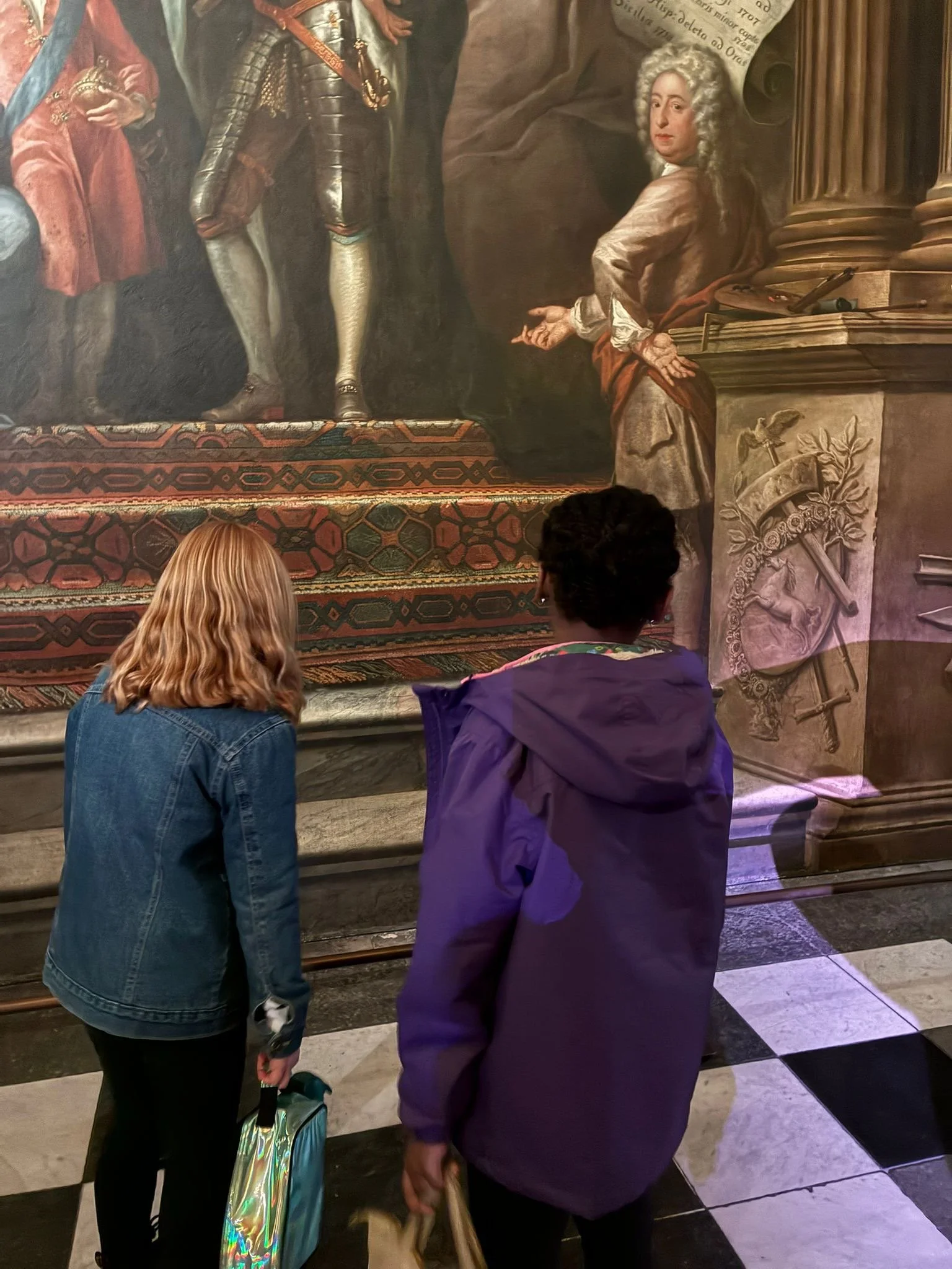 Two women with backpacks looking at a classical painting of a historical figure on a large canvas in a museum.