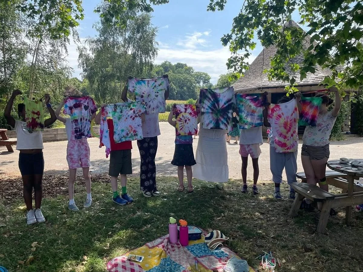 People hanging tie-dye shirts on a line outdoors under trees in a park or backyard on a sunny day.