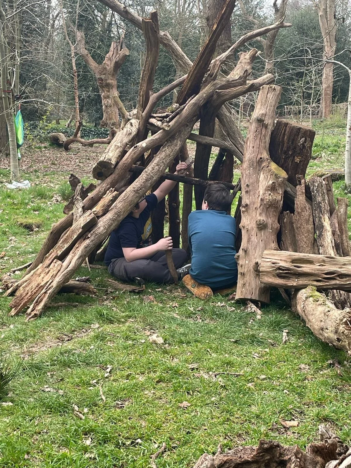 Two children sit inside a small shelter made of large, weathered tree logs and branches in a wooded outdoor area, engaging in an activity together.