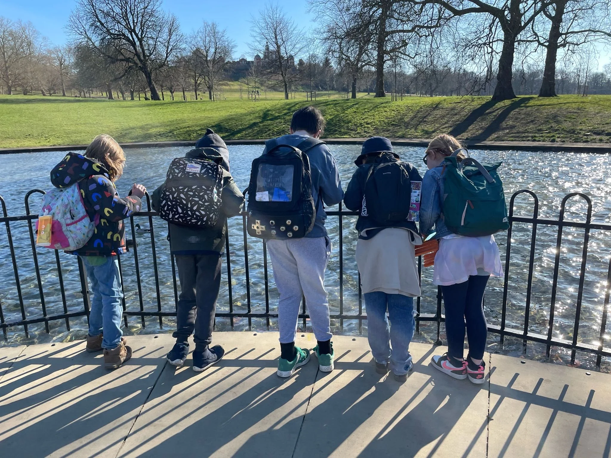 Group of six children with backpacks looking at a pond in a park on a sunny day.