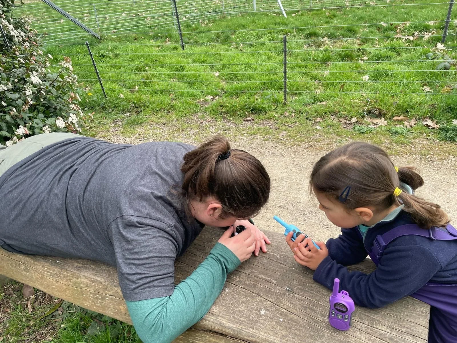 A woman and a girl are lying on a wooden bench outdoors, closely examining a small object with a magnifying glass while holding a walkie-talkie, in a grassy area with a wire fence in the background.
