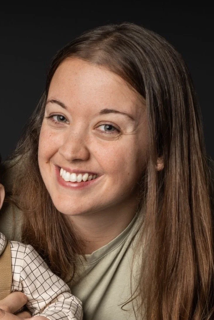 Close-up of a young woman with long brown hair smiling, showing her teeth, against a black background.