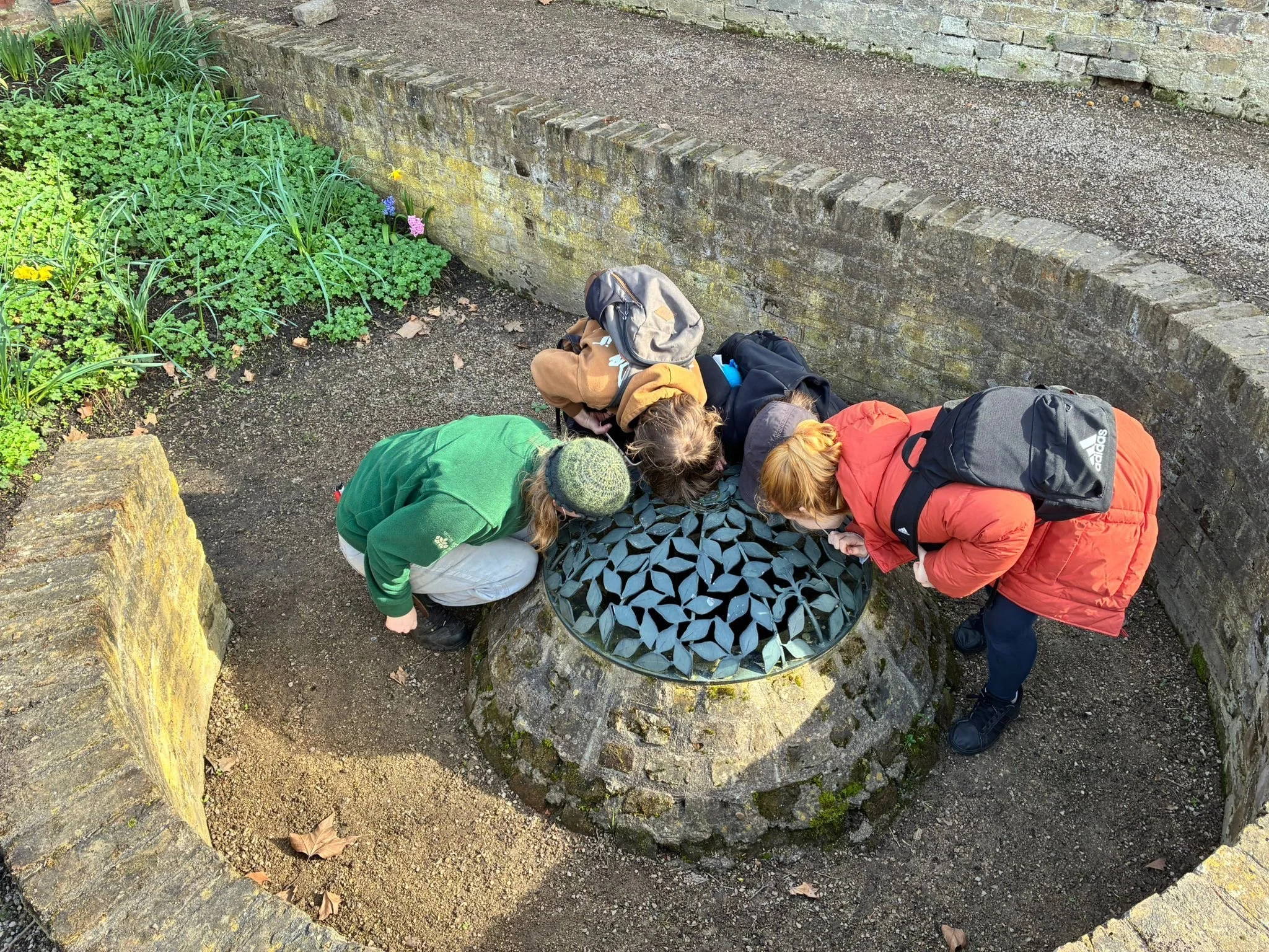 Four people gathered around a small circular mosaic table in a garden, looking at something on the table. They are dressed in warm clothing, with backpacks on their backs. The garden has a flower bed with pink and yellow flowers, and a stone wall sur