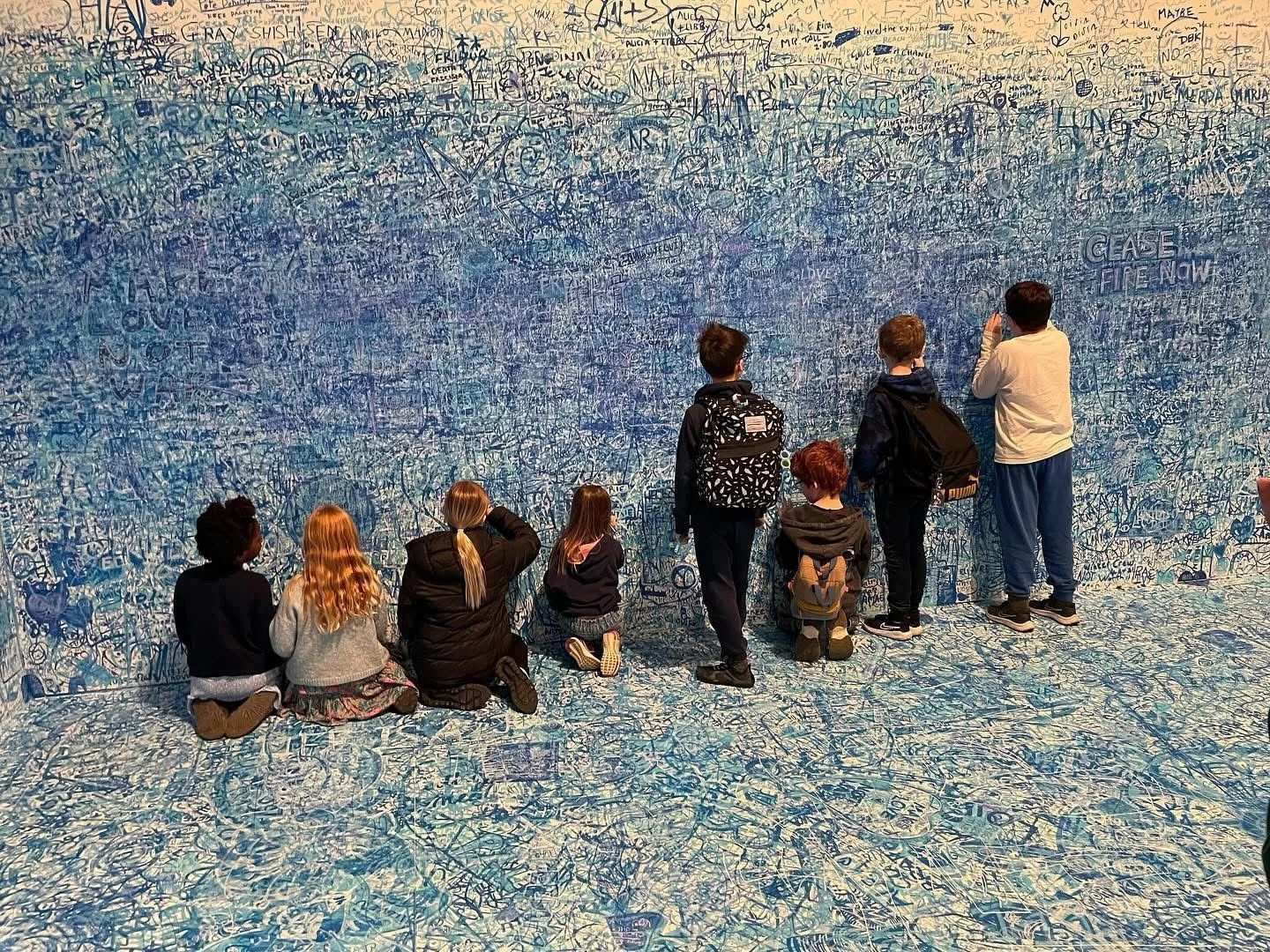 Group of children at an art exhibit, sitting and standing in front of a wall covered with blue graffiti-style drawings and writings, some kneeling, others standing and taking photos.