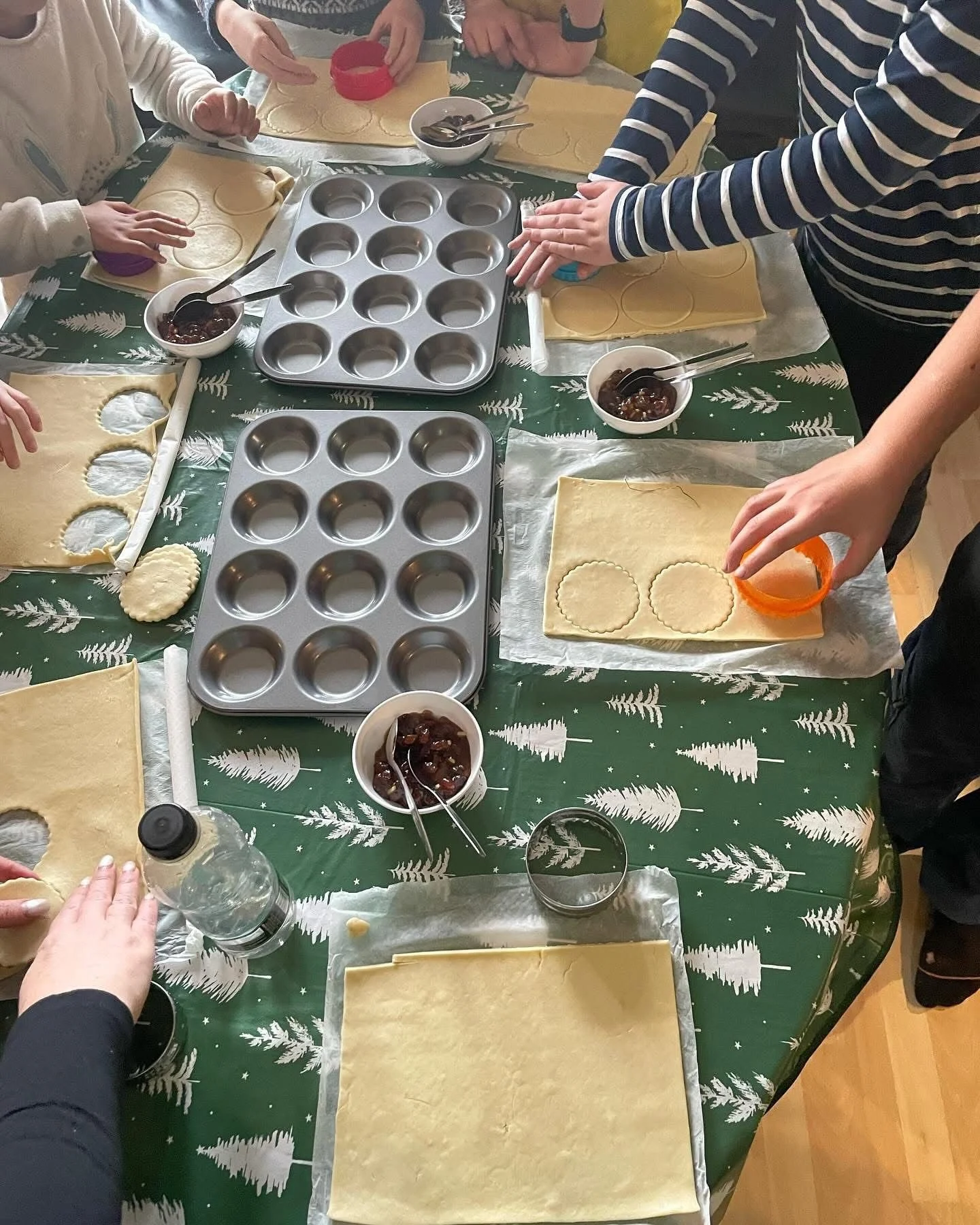 People making holiday cookies with rolled out dough, cutters, and bowls of jam on a festive table.