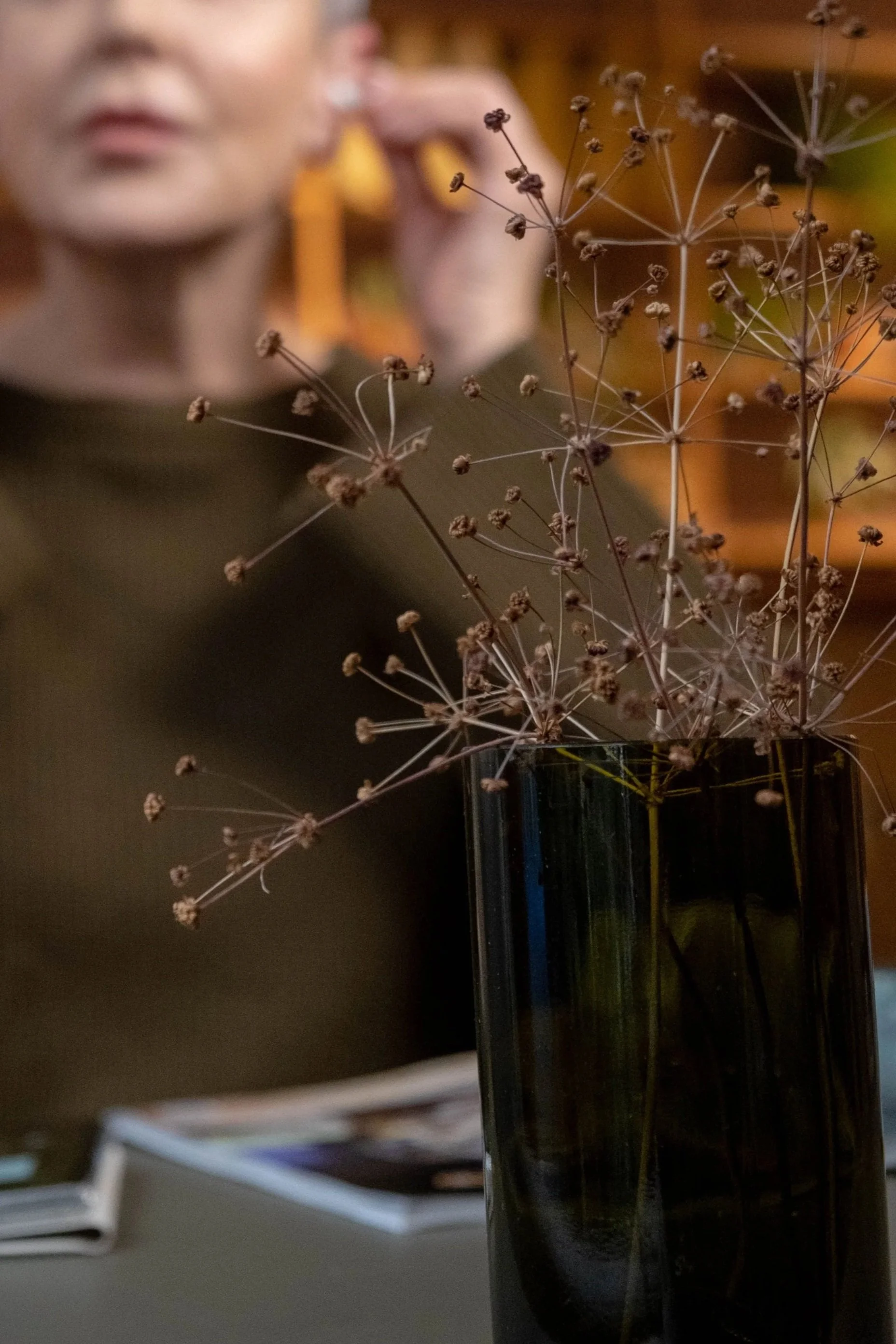 Close-up of dried brown plant with small round seed heads in a black glass vase, blurred background.