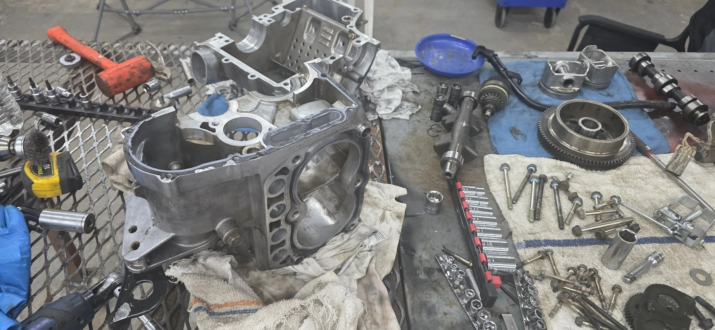 A workshop table with disassembled engine parts, tools, bolts, and gears, indicating automotive repair activity.