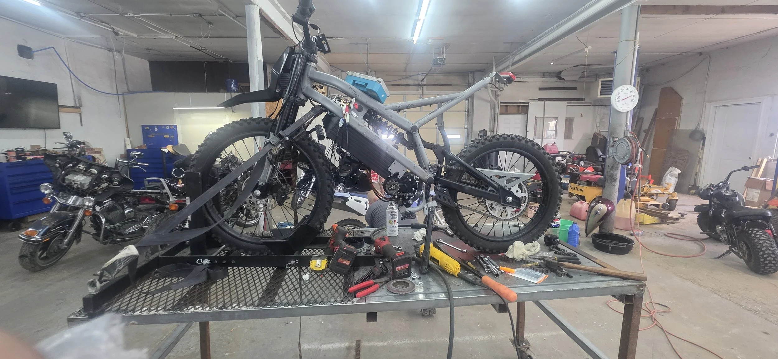 A motorcycle in the process of being built or repaired on a metal worktable in a workshop surrounded by tools, parts, and other motorcycles.