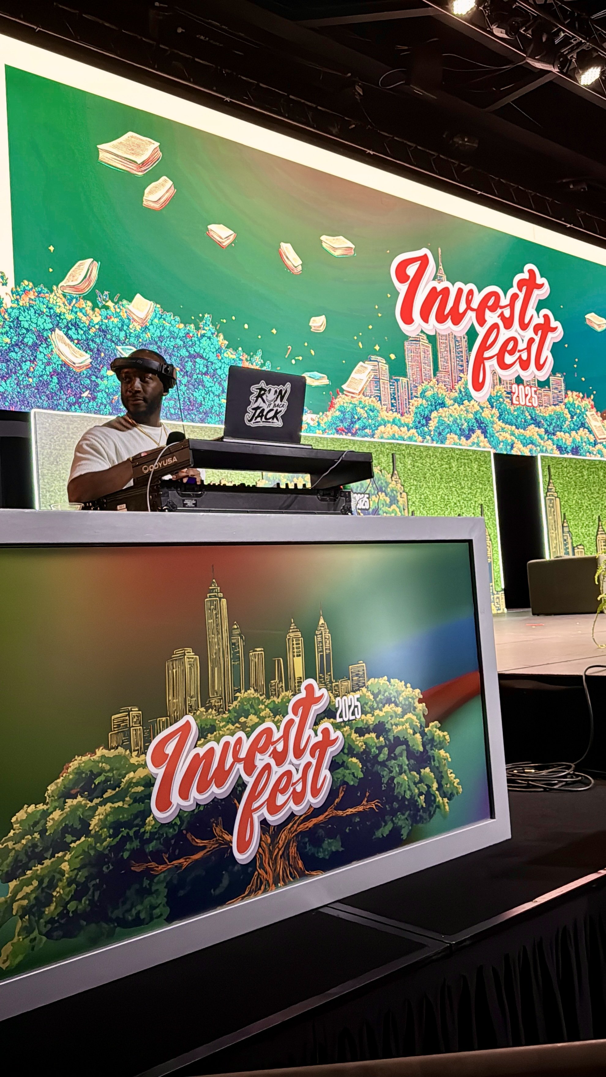A DJ performs on stage at Invert Fest 2025, with large colorful digital screens displaying a cityscape, books, and the event logo in the background.