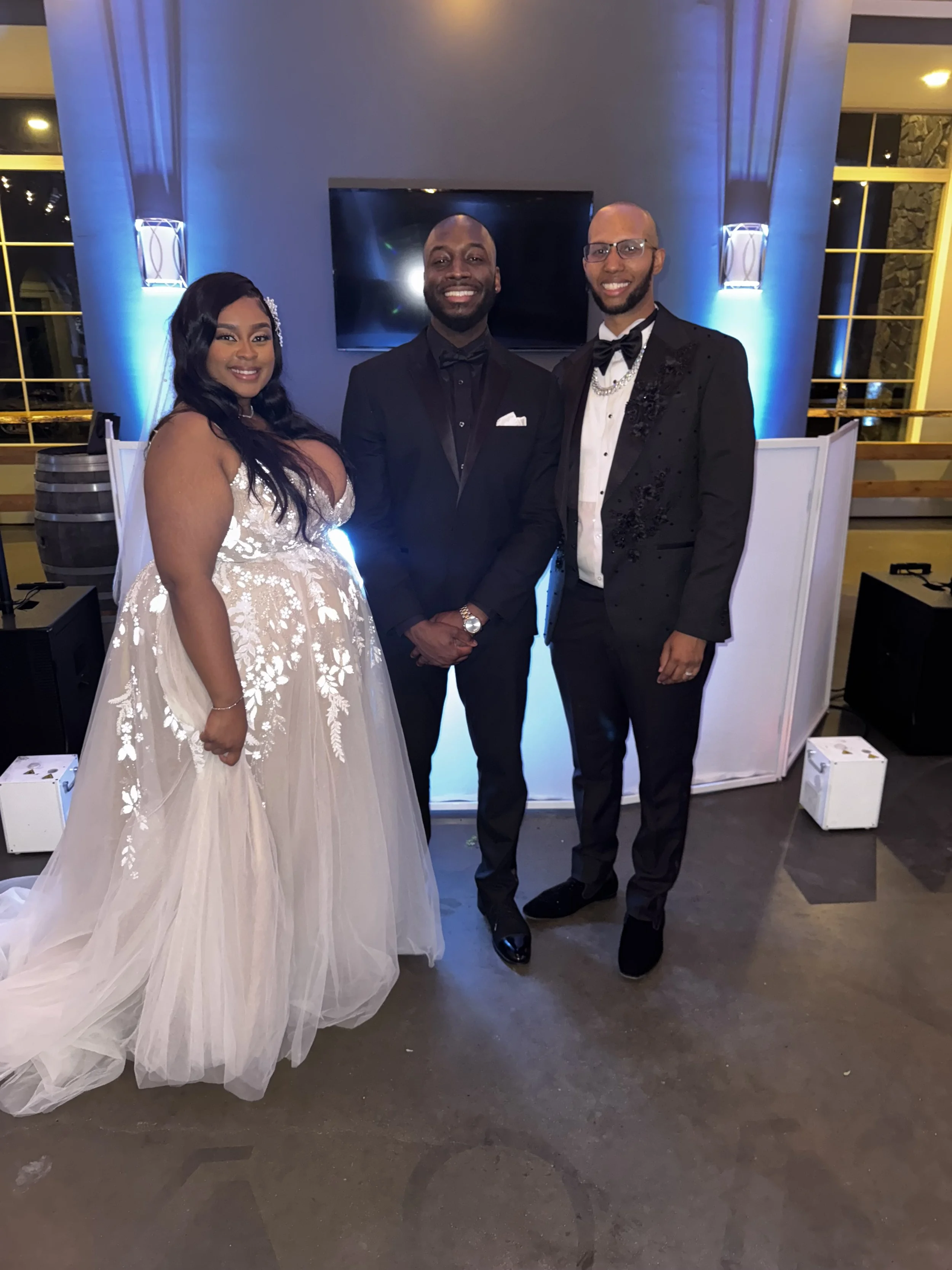 Three people in formal attire standing together at a celebration or wedding. The woman on the left is wearing a white gown with floral embroidery, and the two men are in black tuxedos. They are smiling and posing indoors with blue lighting and large 