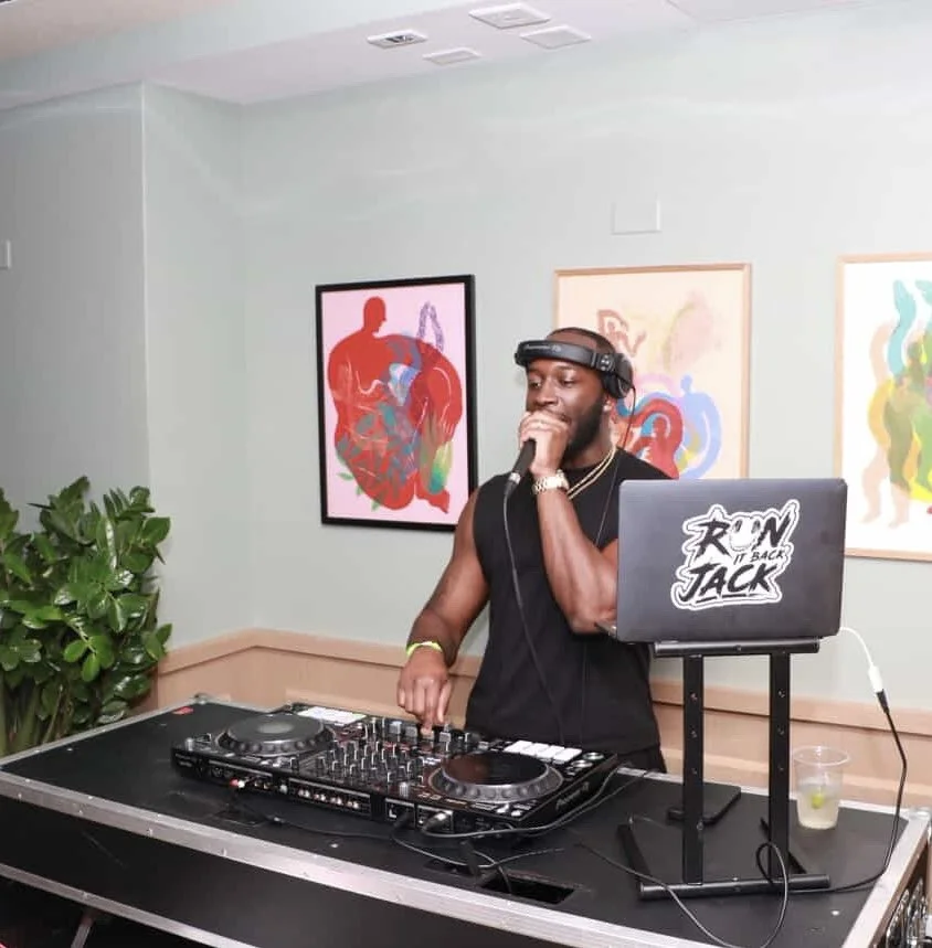 A man wearing a black sleeveless shirt, headphones, and a watch, standing behind a DJ controller, speaking or singing into a microphone, with abstract art paintings on the wall behind him.