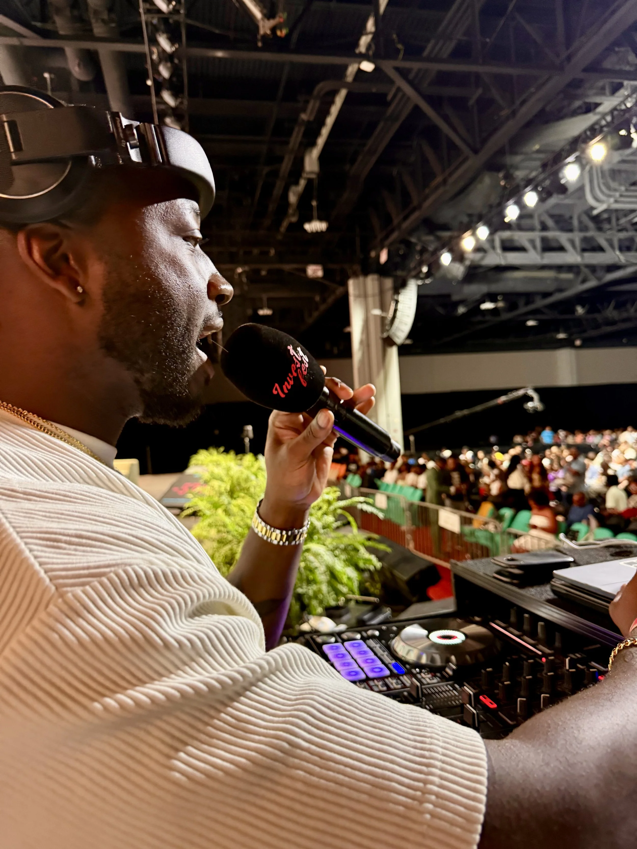 A DJ or radio host speaking into a microphone with an audience in the background at an indoor event.