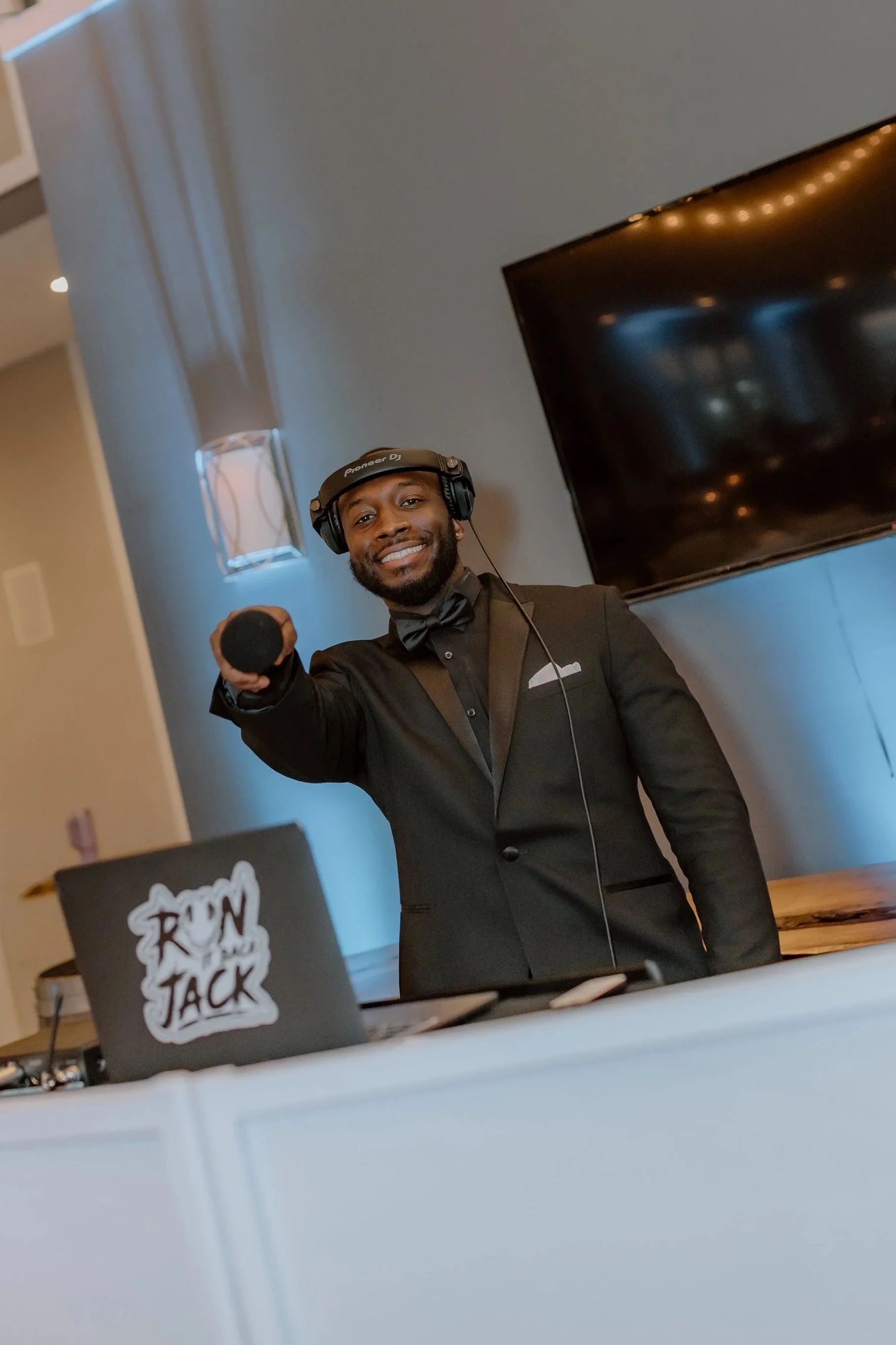 Male DJ in black tuxedo with bow tie, wearing headphones, smiling and holding a microphone, standing behind a DJ booth.