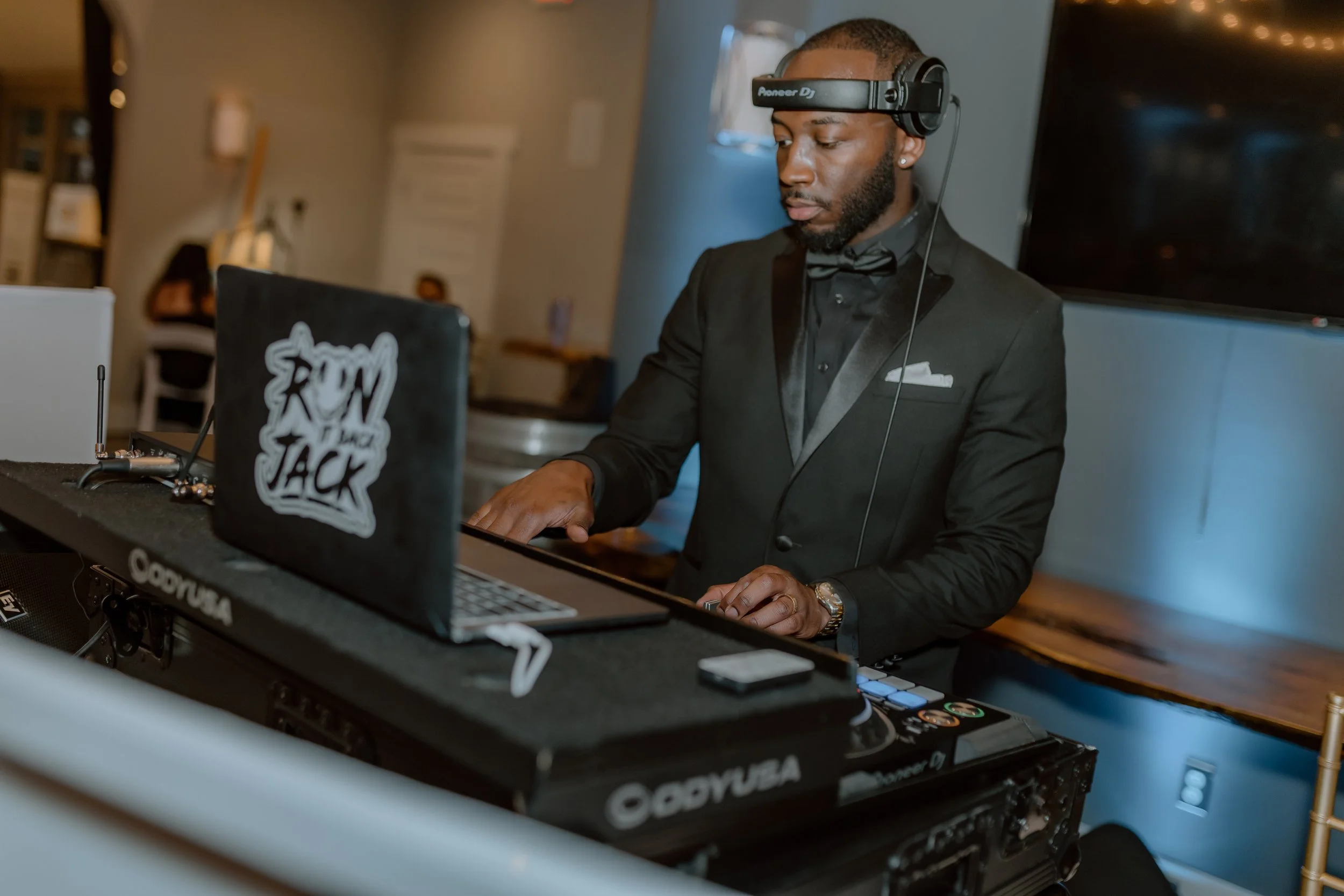 A man dressed in a black tuxedo with a bow tie, wearing headphones, DJing at an event with a laptop and DJ equipment.