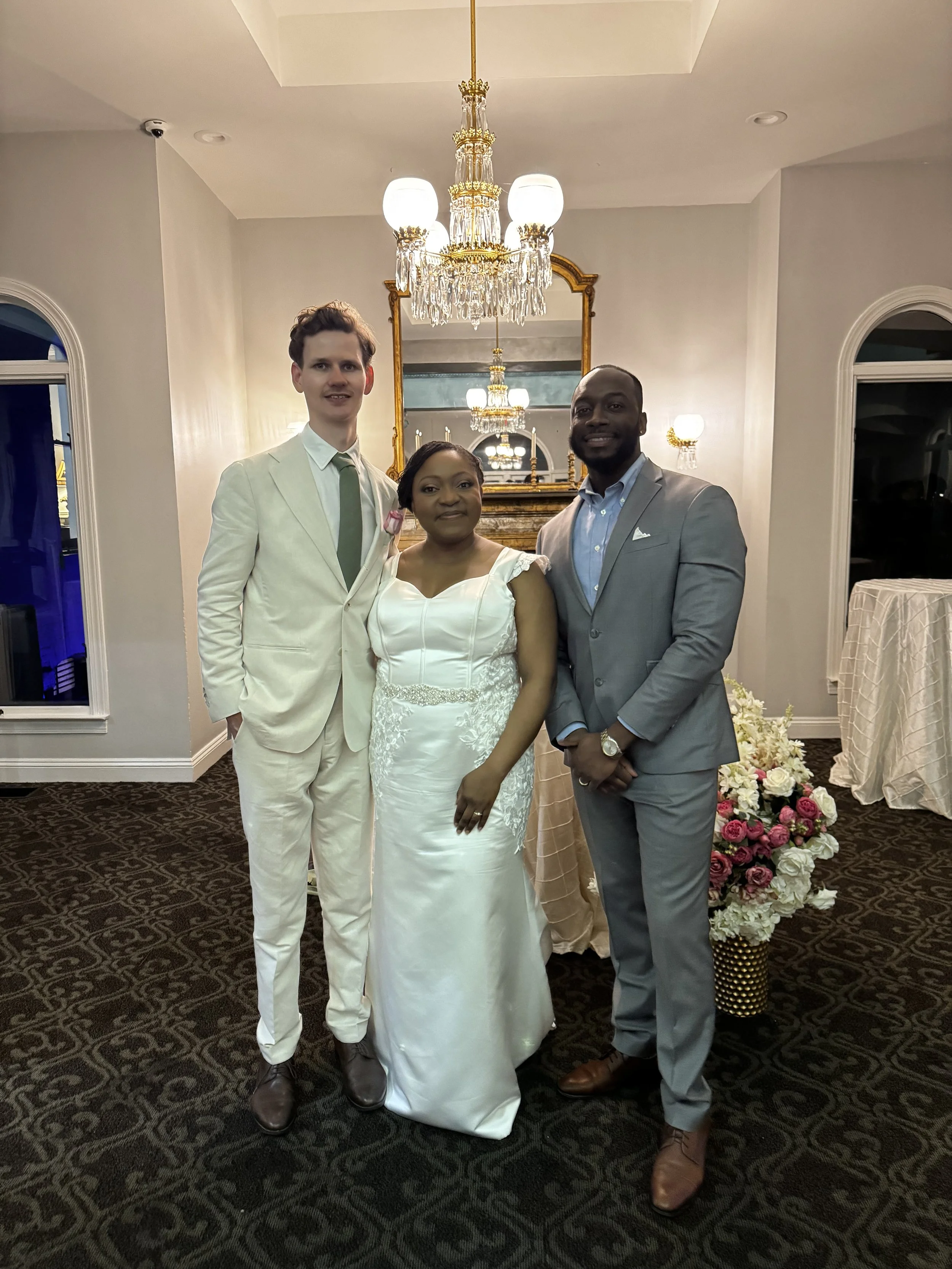 Three people dressed in formal attire standing together in a decorated room with chandeliers, mirror, and floral arrangements.