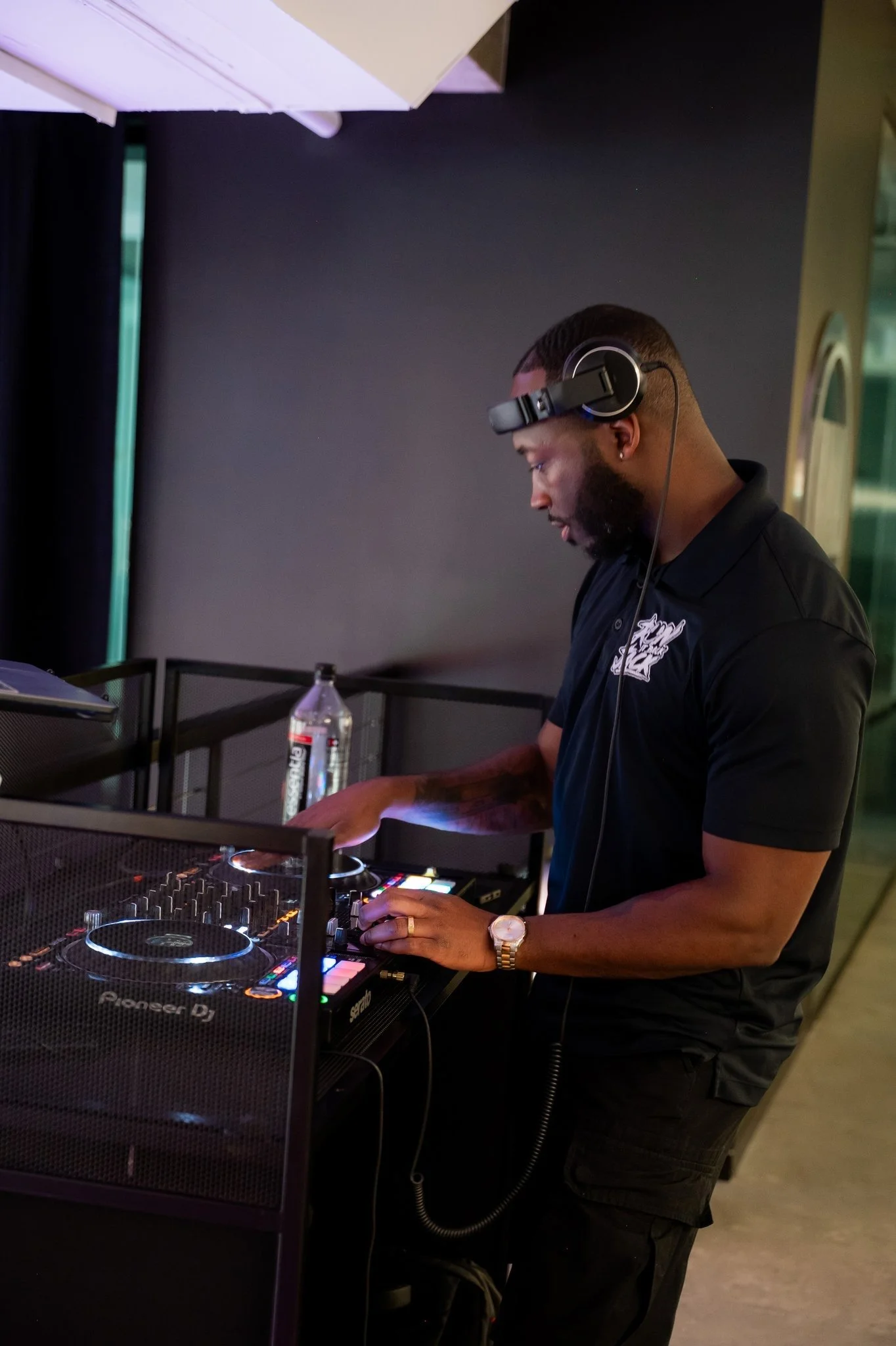 A DJ wearing headphones and a black polo shirt operating a Pioneer DJ controller in a dimly lit venue.