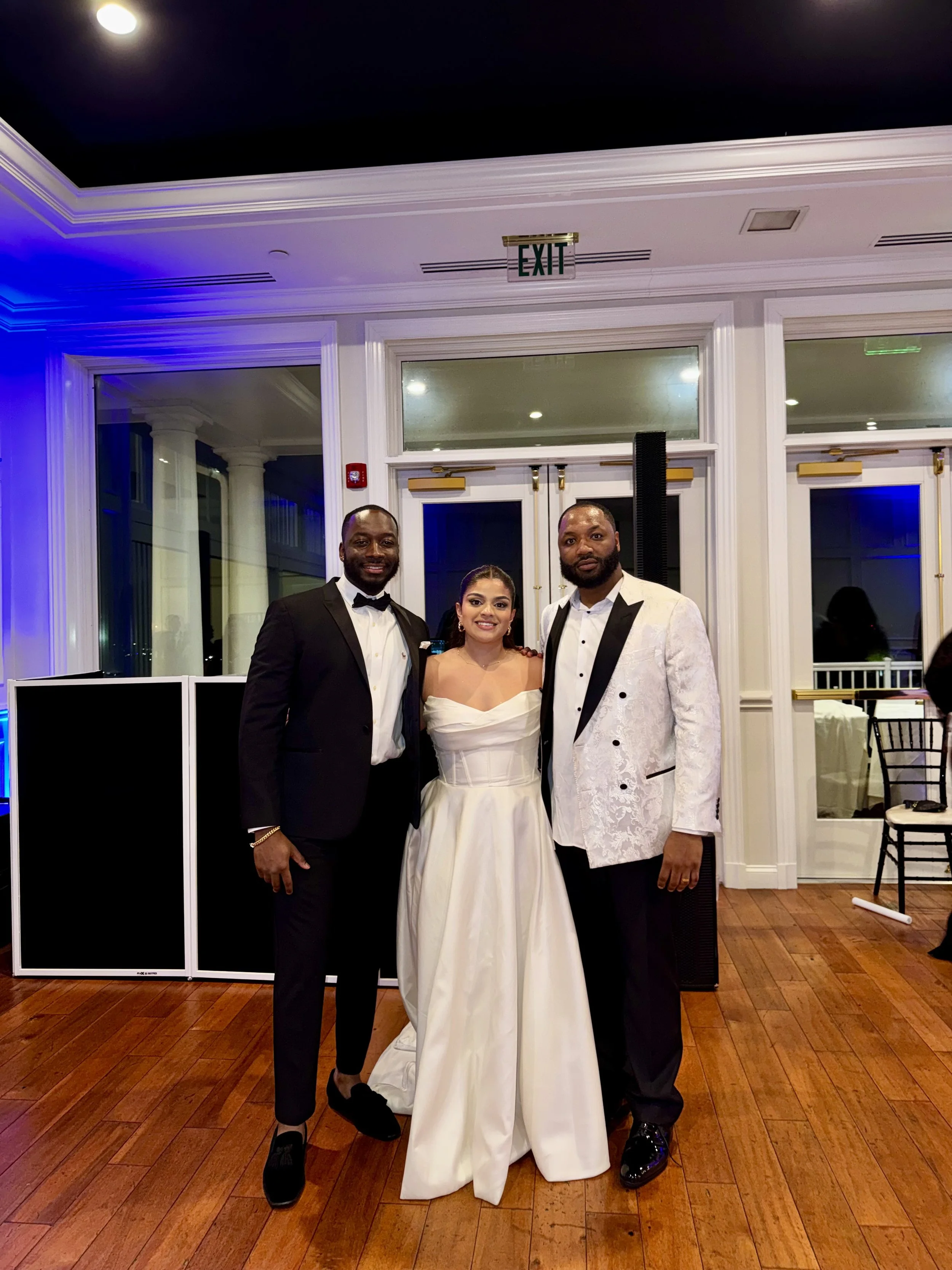 Three people in formal attire standing together indoors at a wedding reception. The woman in the center wears a white wedding dress, and the two men on either side wear tuxedos.