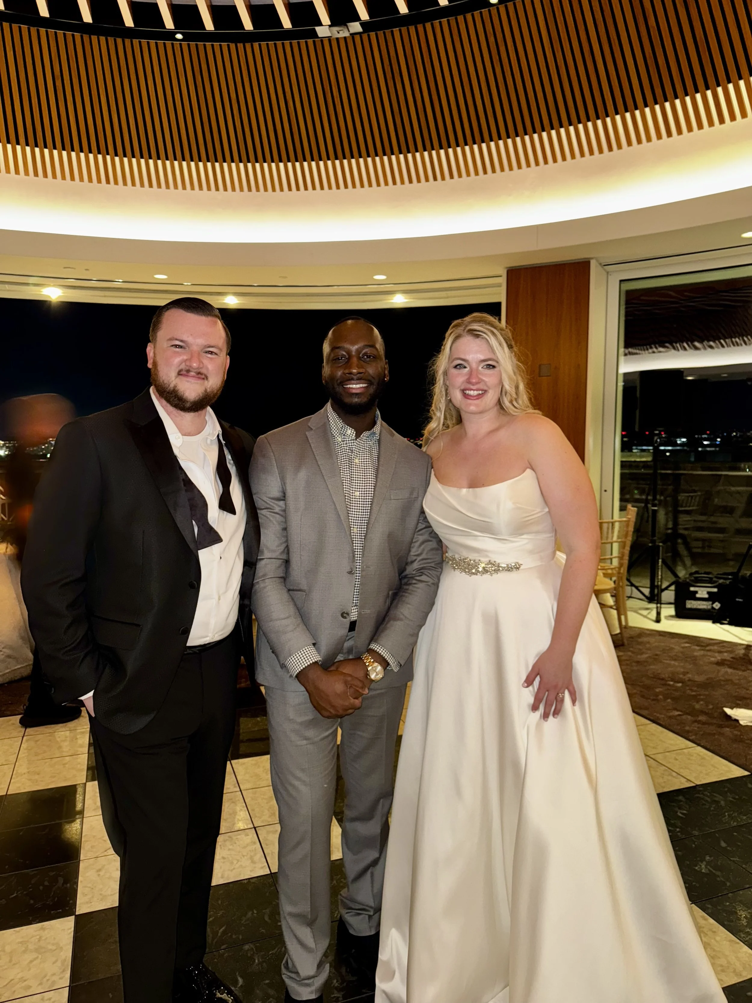 Three people dressed in formal attire standing together in a venue with a wooden ceiling and city lights visible through a large window.