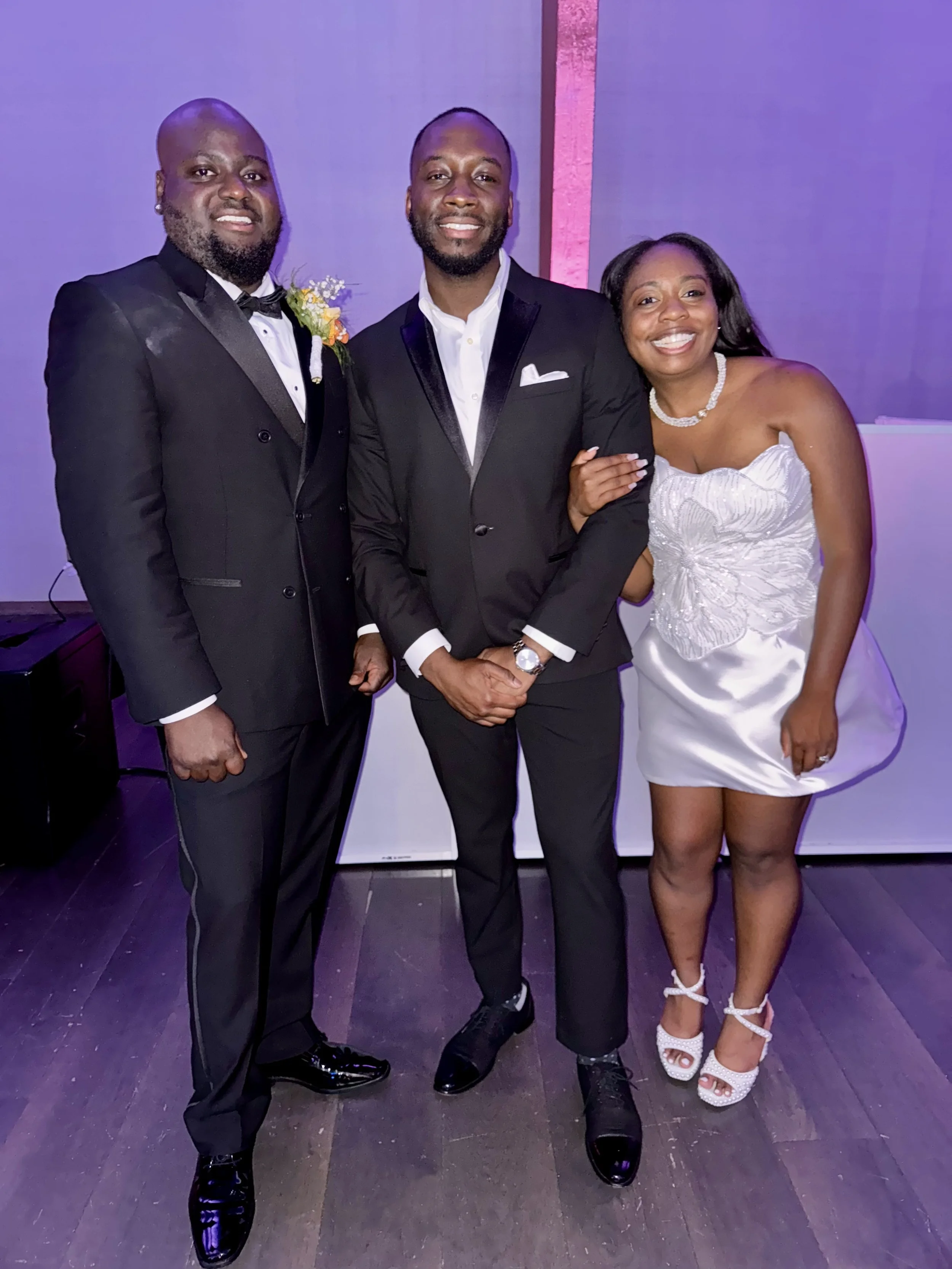 Three people dressed in formal attire at an event, standing together and smiling for the camera. The two men are in black tuxedos, and the woman is in a white dress with high-heeled shoes.
