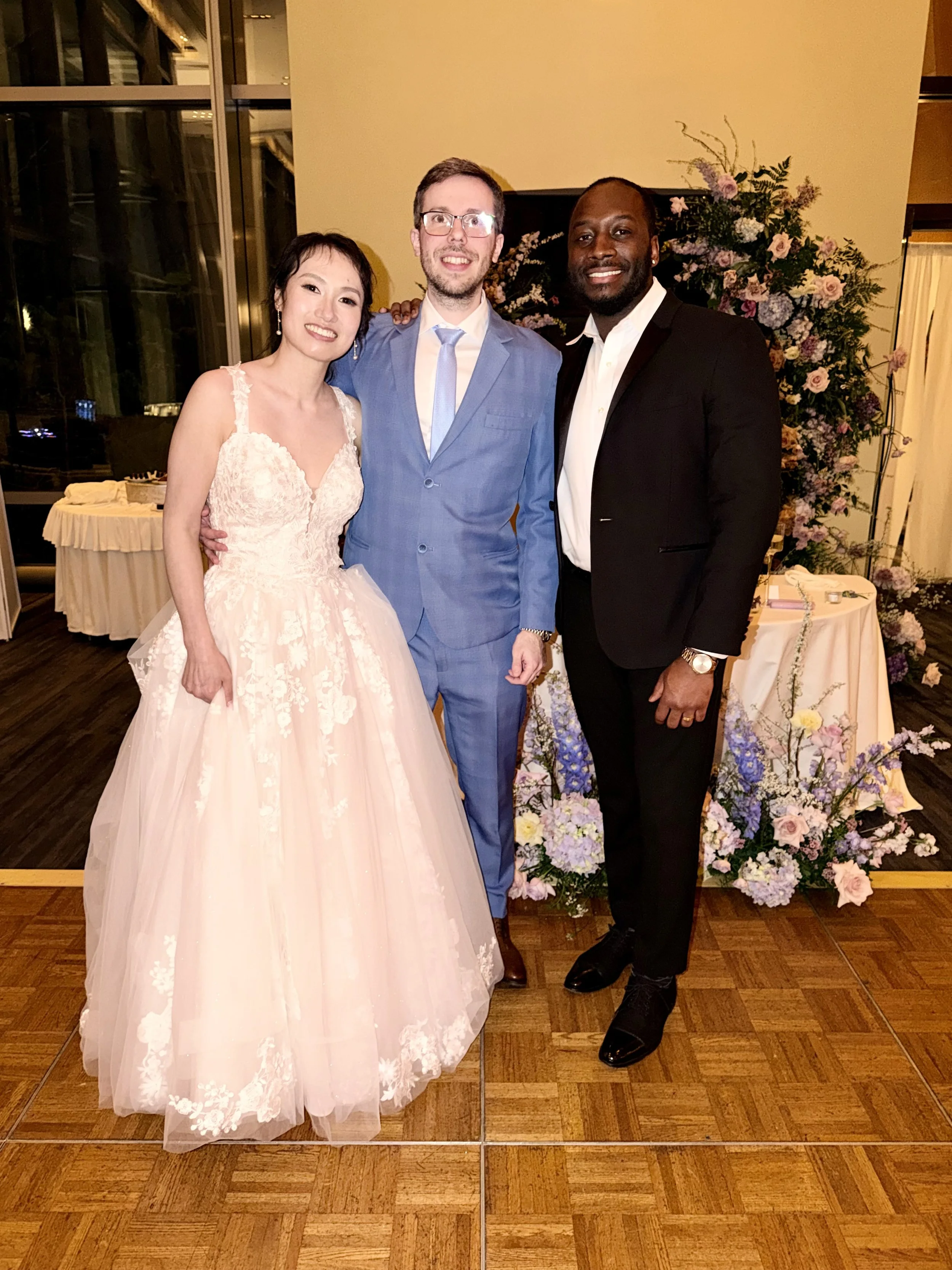 A group of three people dressed in formal attire at a wedding reception, standing on a wooden floor in front of a floral backdrop.