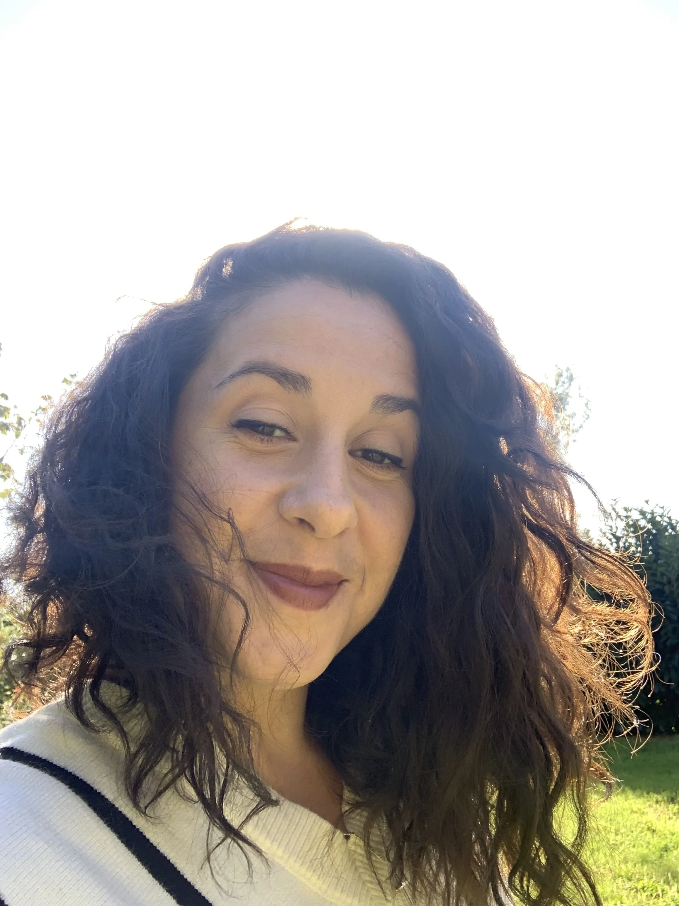Une femme (Kalie de Mystencia) avec des cheveux bouclés noirs, souriant légèrement, en extérieur en pleine lumière du soleil, portant un pull blanc avec des rayures noires.