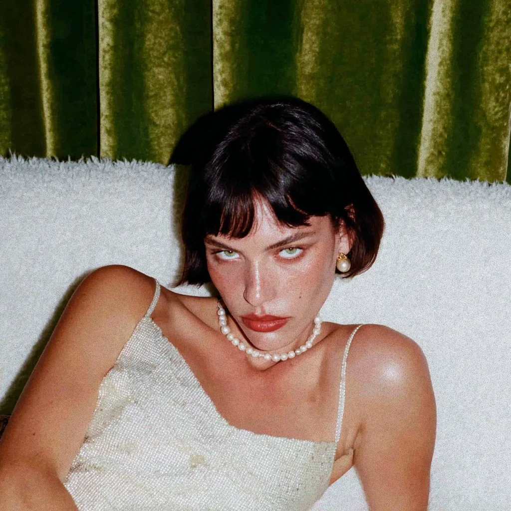 A woman with short black hair, wearing pearl jewelry and a white, beaded dress, is reclining on a white surface with green curtains in the background.