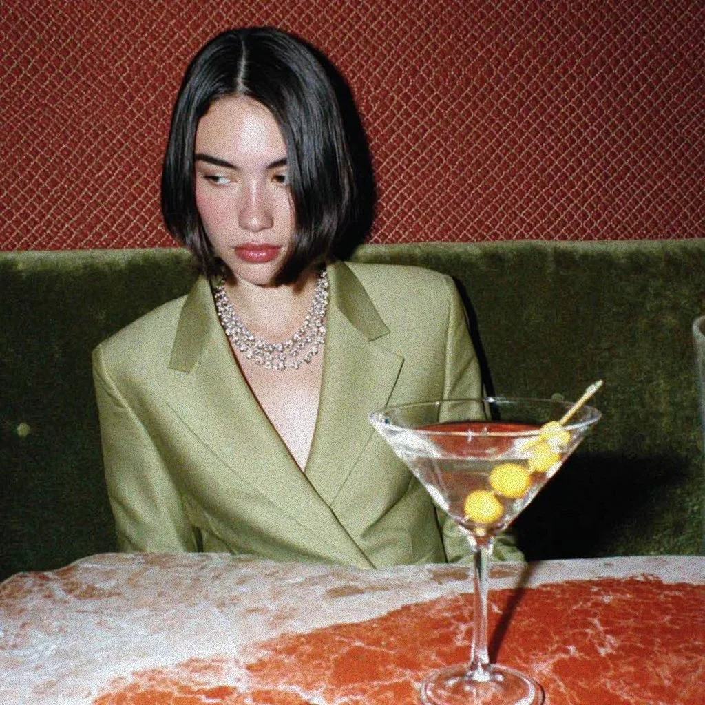 A woman with black hair wearing a green blazer and a pearl necklace sitting at a table with a large martini glass filled with a clear beverage, garnished with olives and a stir stick.