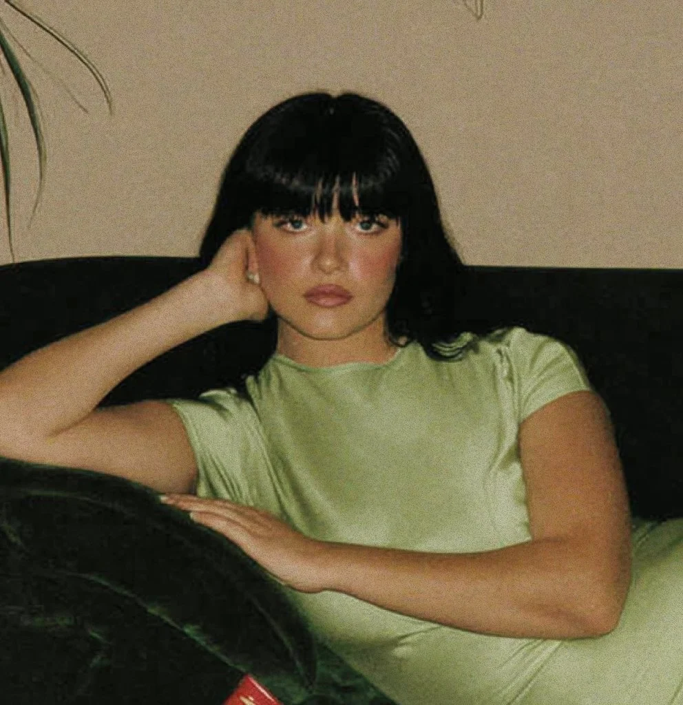 A woman with dark hair and bangs sitting on a black sofa, wearing a light green T-shirt, resting her head on her left hand and looking at the camera.