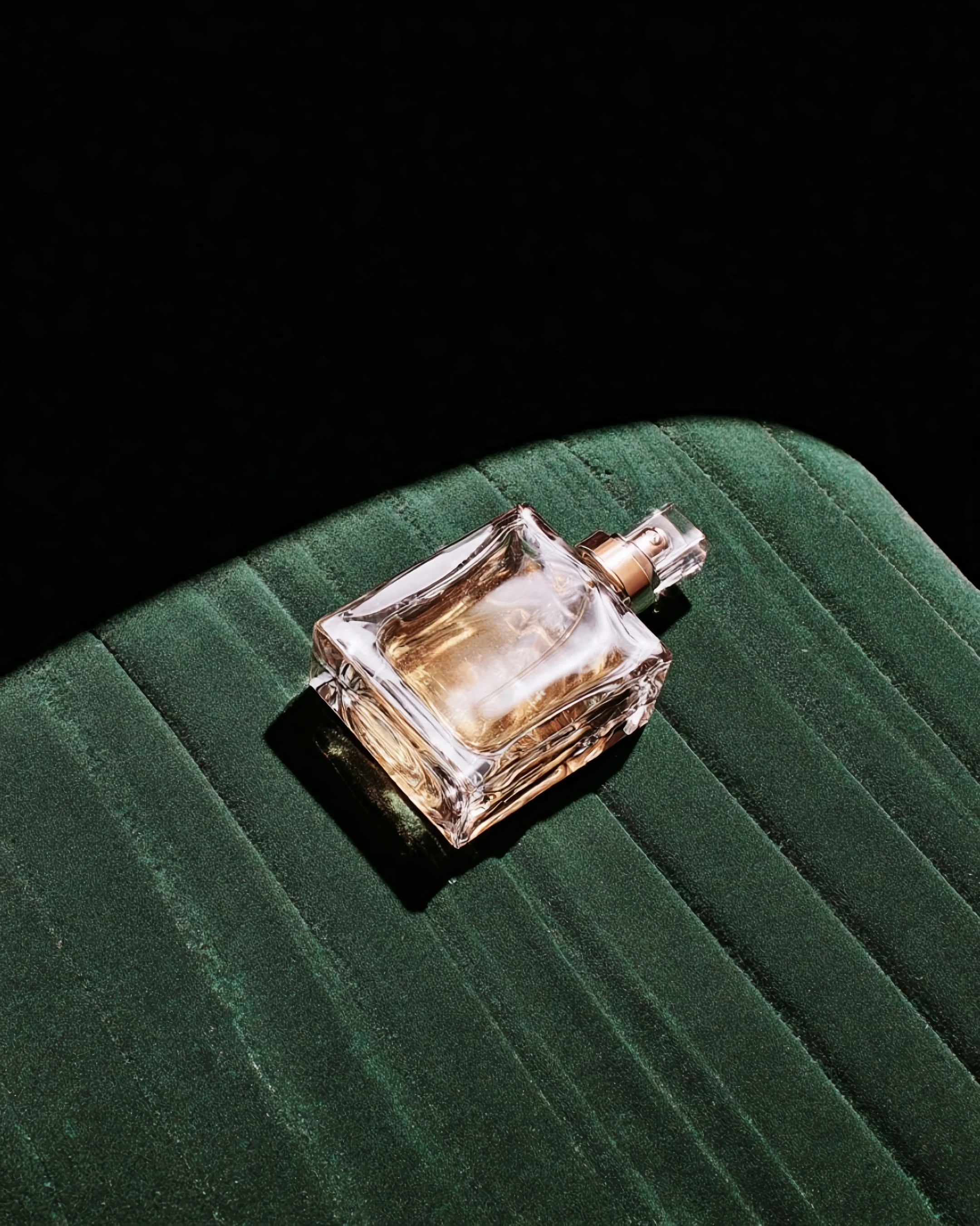 A clear glass perfume bottle with a transparent stopper, lying on a green textured surface, with a dark background.