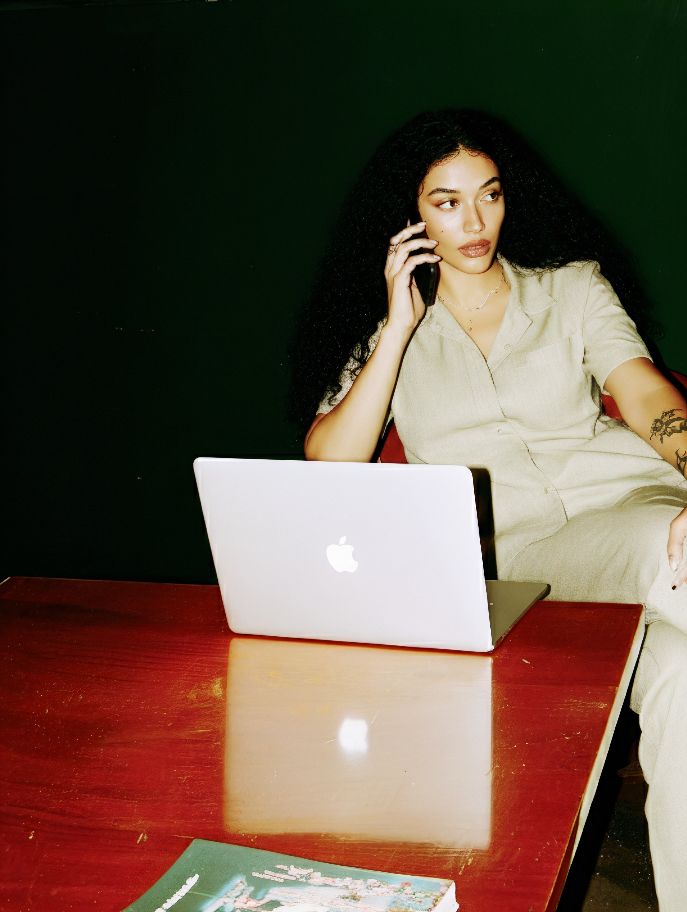 A woman with curly black hair sitting at a wooden table, talking on her cell phone with a MacBook laptop in front of her and a magazine on the table.