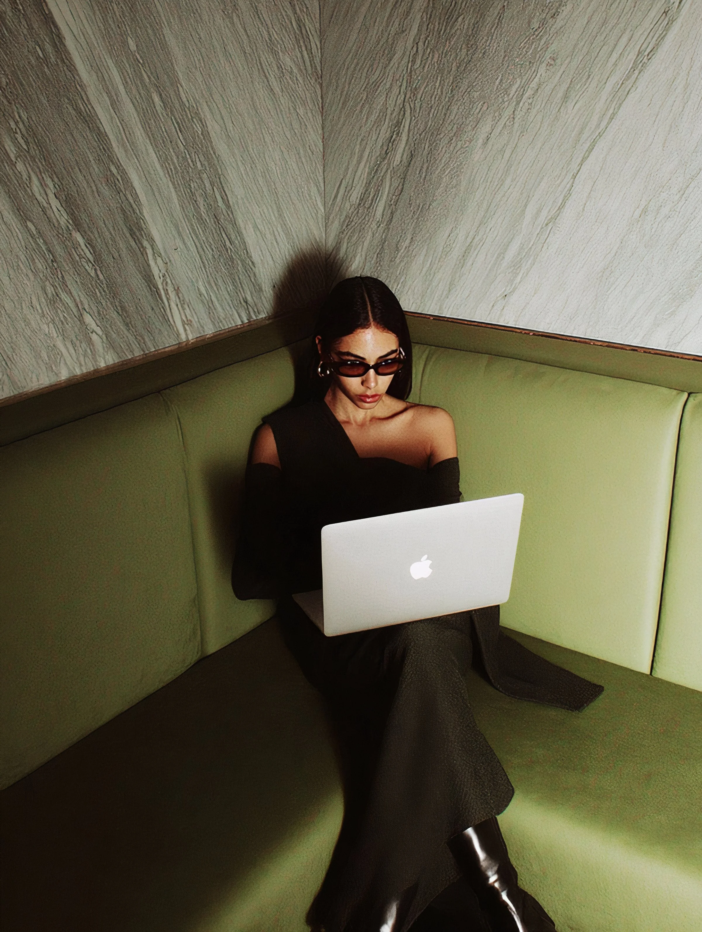 A woman with dark hair, wearing sunglasses and black clothing, sitting on a green booth seat in a booth, using a silver laptop with a white Apple logo, with her legs crossed and wearing shiny black boots, against a wooden and textured wall background.